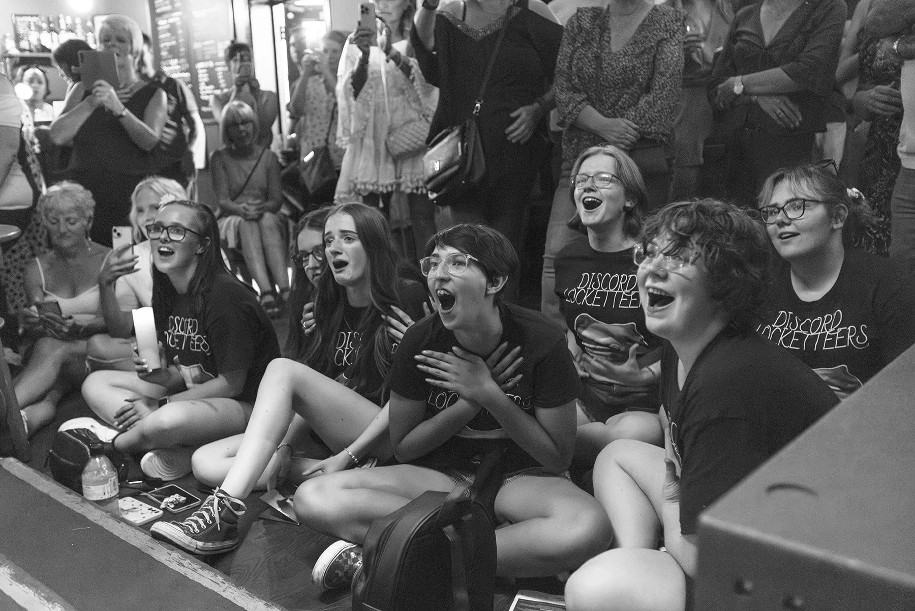 A group of young women and some older women are sitting and watching an event with excitement and joy. They are wearing matching black T-shirts with the 'Discord Rockettes' logo. Some are holding phones and one woman appears to be emotional with her 
