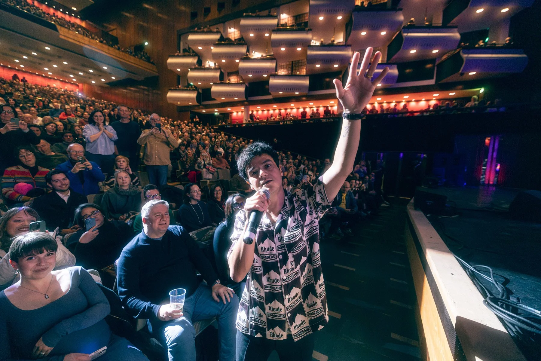 Jamie Cullum holding a microphone and waving to the audience during a comedy or entertainment event in a theater or concert hall filled with seated spectators watching and smiling.