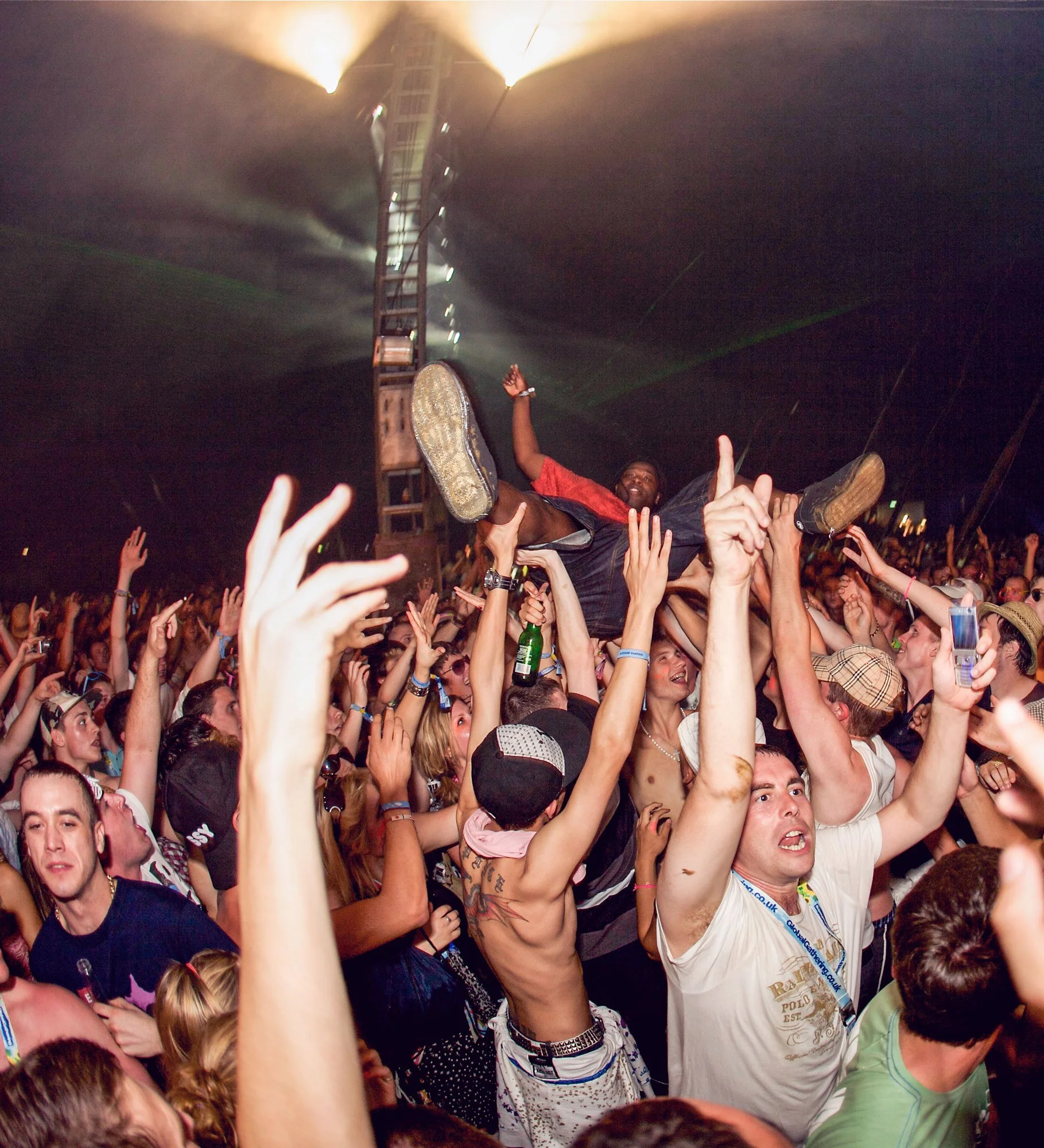 Festival crowd with a man crowd surfing, surrounded by people reaching up with their hands. The man is smiling, wearing a red shirt and shorts, and is being held up by the crowd. Stage lights are visible at the top of the image.