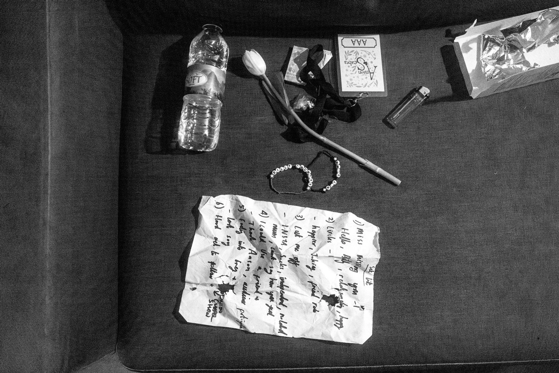 A desk with two water bottles, a tulip flower, a small box labeled 'Anson Sebera', a lighter, a crumpled handwritten set list, a beaded bracelet.