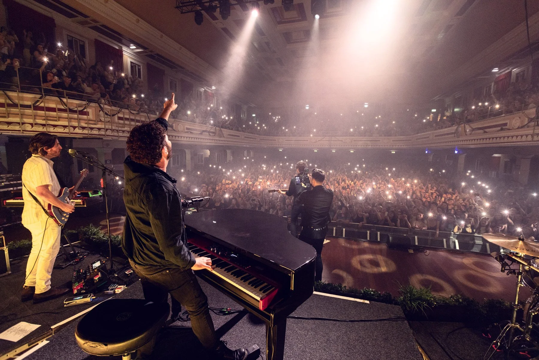 Dean lewis and Billy Lockett performing on a stage with a grand piano, guitar, and keyboard in front of a large audience. Audience members are holding up their phones and lights, creating a sea of lights in the darkened theater.
