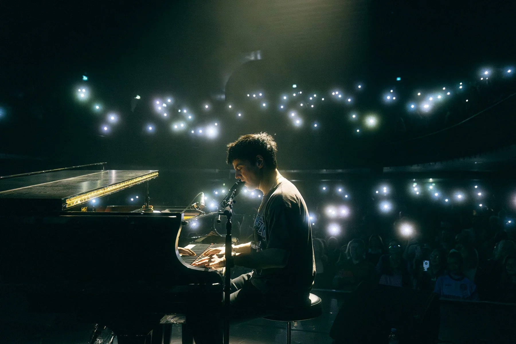 Jamie Cullum playing a grand piano and singing into a microphone on stage, with a dark background filled with small, bright lights, in front of an audience.