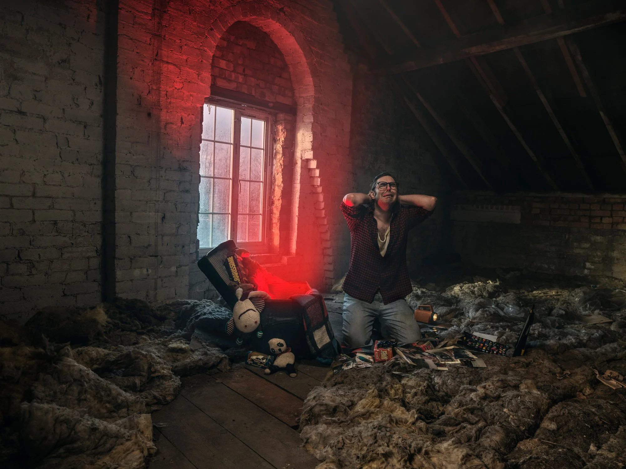 A man with glasses and a checkered shirt stands inside a dim, unfinished attic with exposed brick walls and a sloped ceiling. He appears distressed, holding his neck with both hands. The attic has scattered items, toys, clothing, and a bag on the floor, with a window letting in natural light and red light illuminating part of the wall.