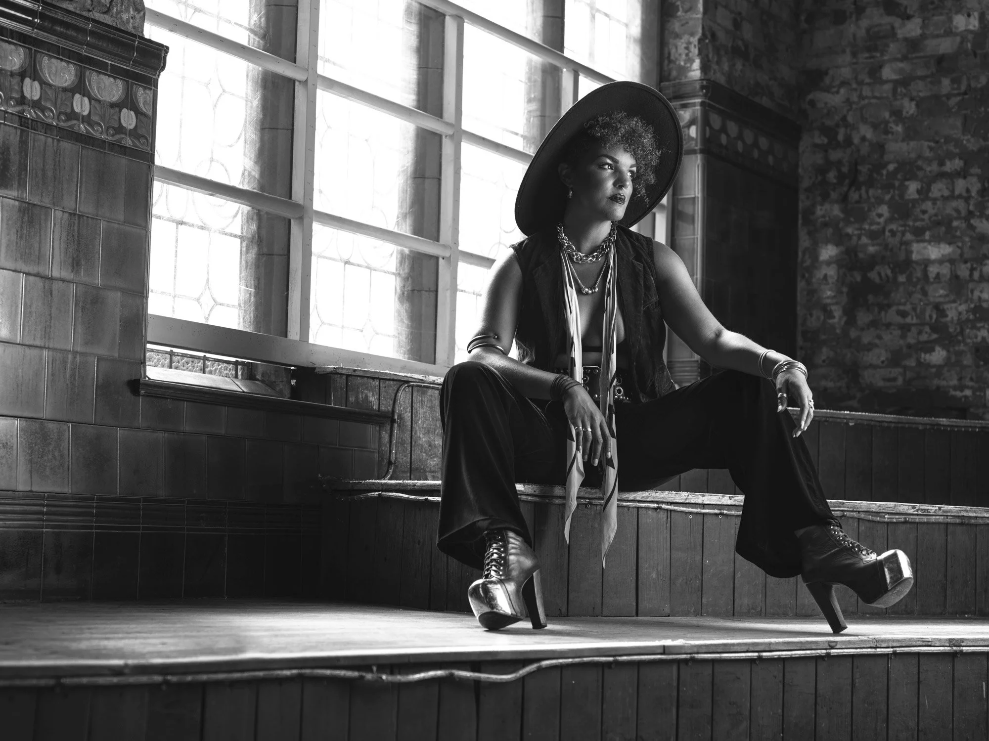 A woman with curly hair wearing a wide-brimmed hat, layered necklaces, vest, and boots, sitting on a staircase in a room with brick walls and large windows.