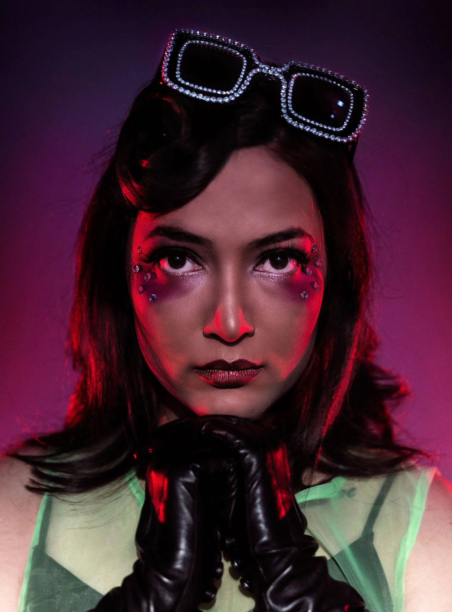A woman with black hair and glittery makeup wearing large, square sunglasses with rhinestones and black leather gloves, in front of a purple-pink background.