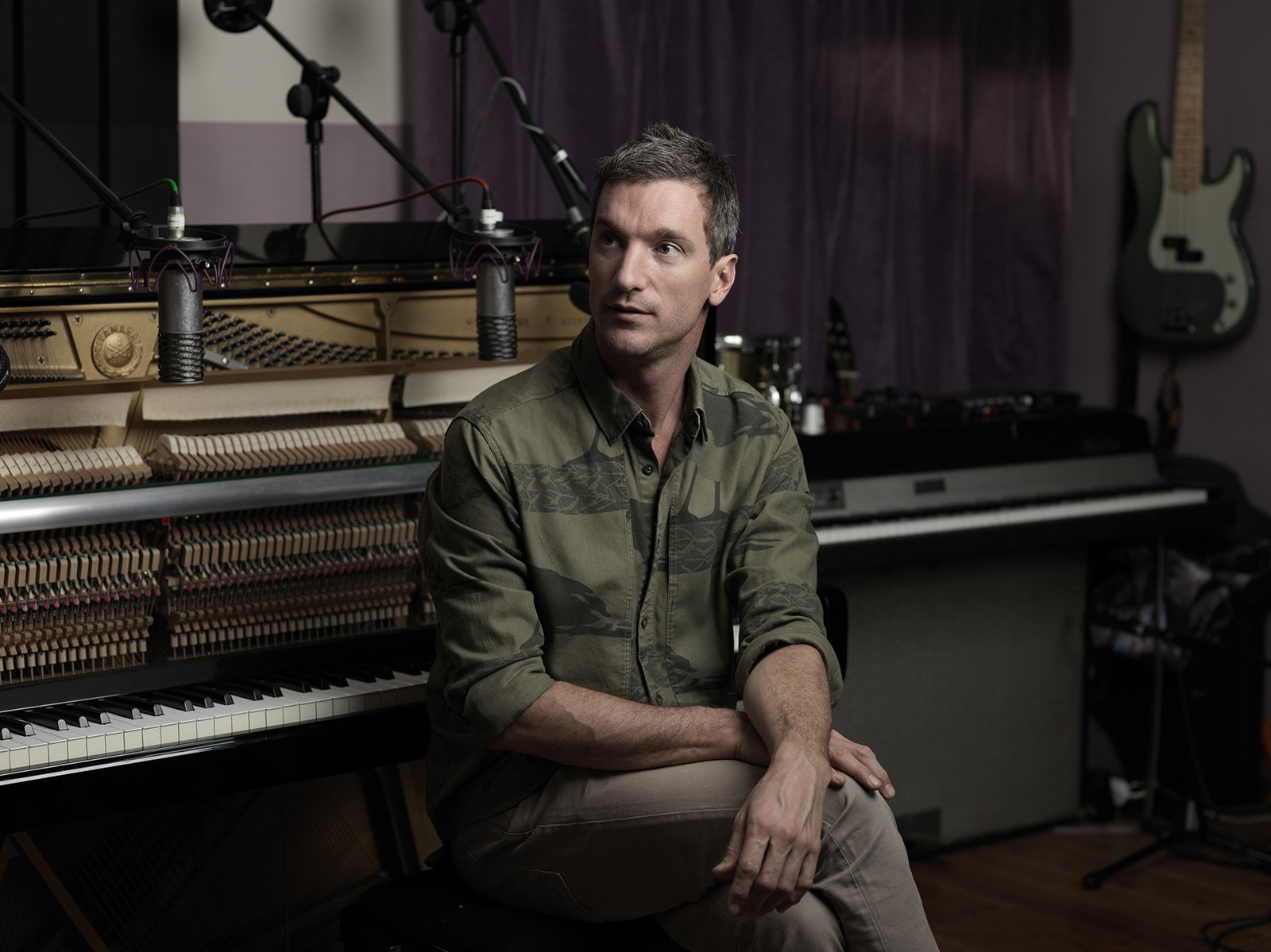 Andy Barlow from Lamb sitting in front of an open piano in a music studio, with microphones set up above the piano, a guitar hanging on the wall, and a keyboard beside him.