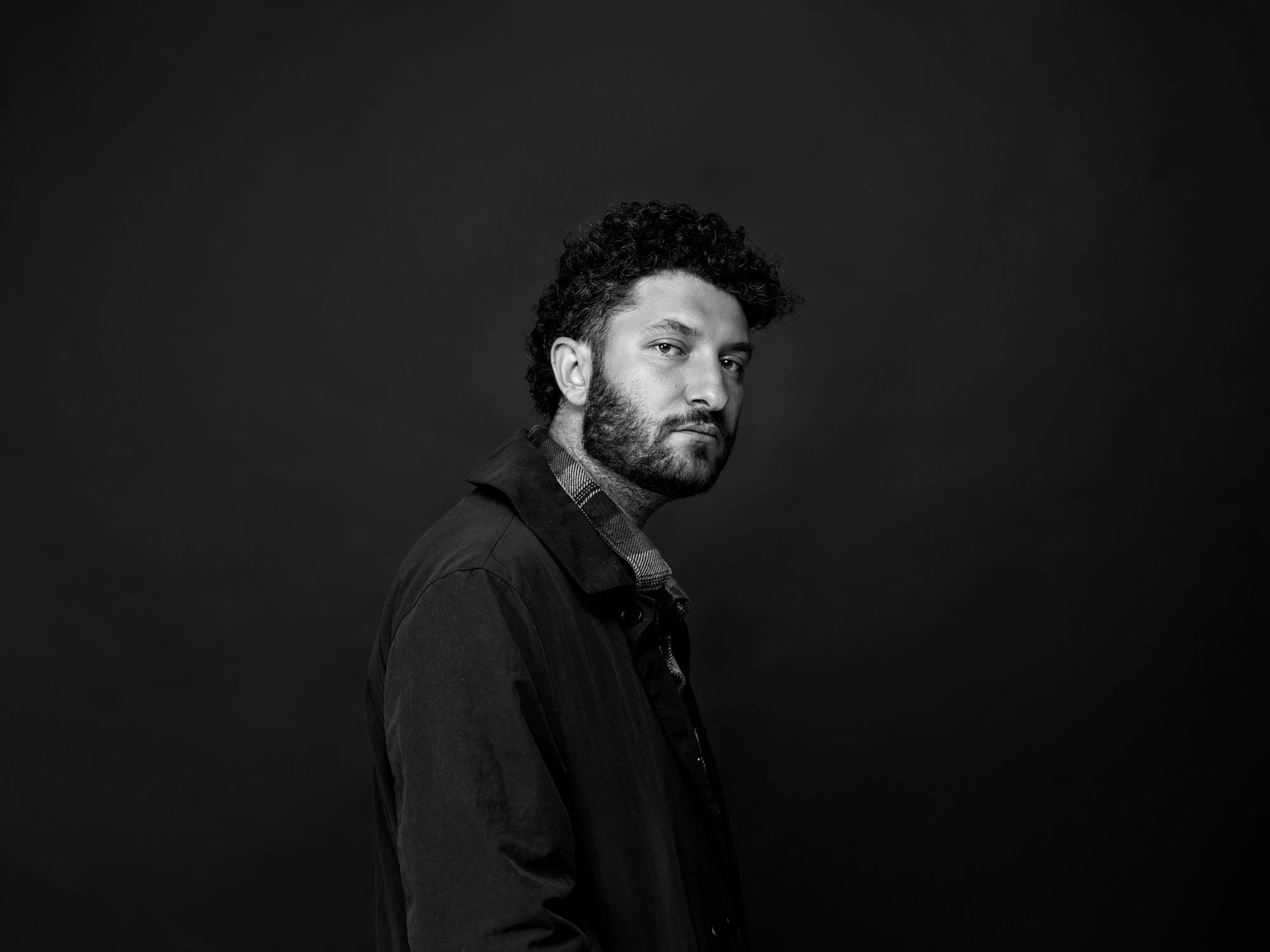 Black and white portrait of a man with curly hair and beard, looking over his shoulder against a dark background.