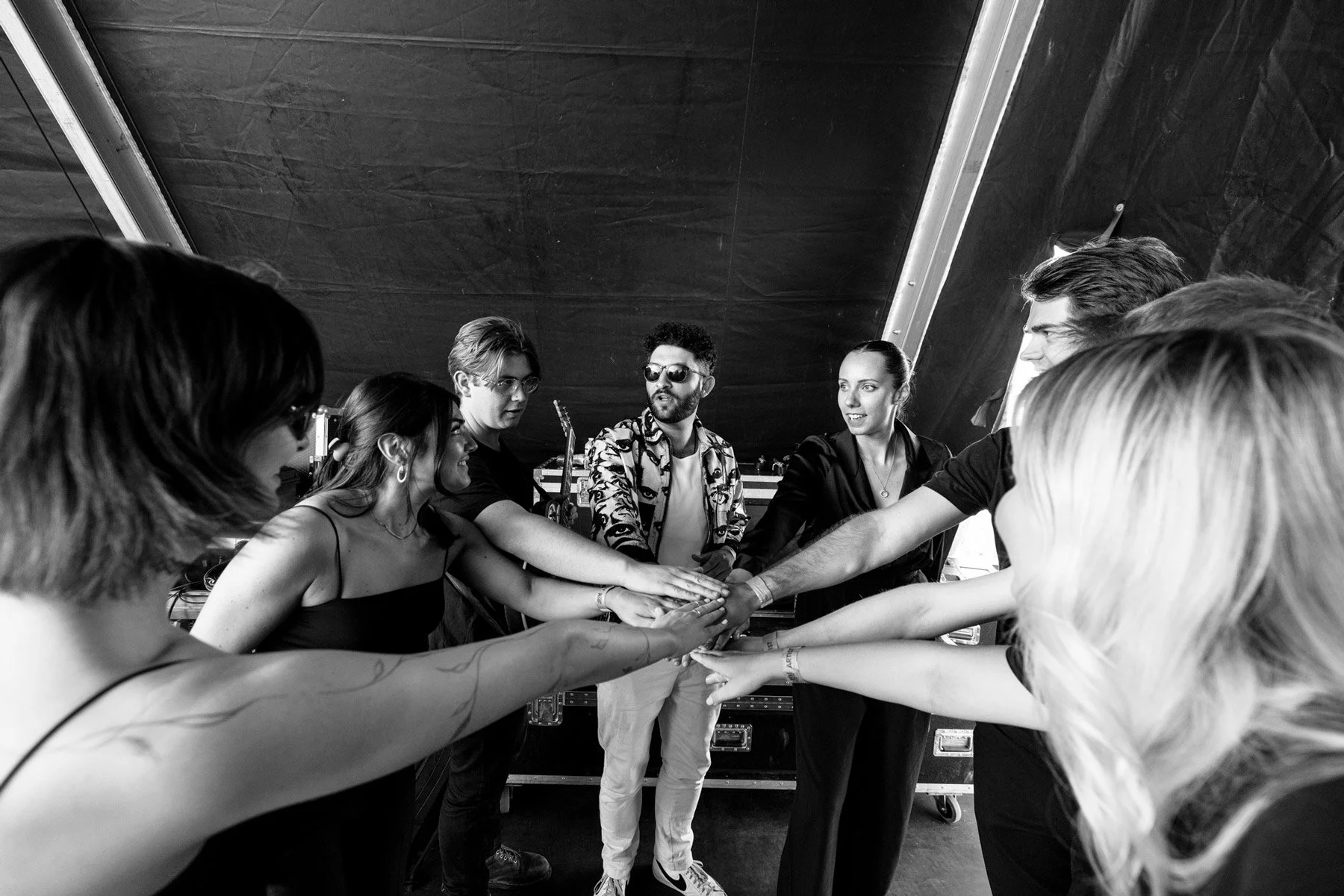 Billy Lockett and band huddled together with their hands stacked in the center, in a collaborative team-building gesture, indoors with a black canopy overhead.