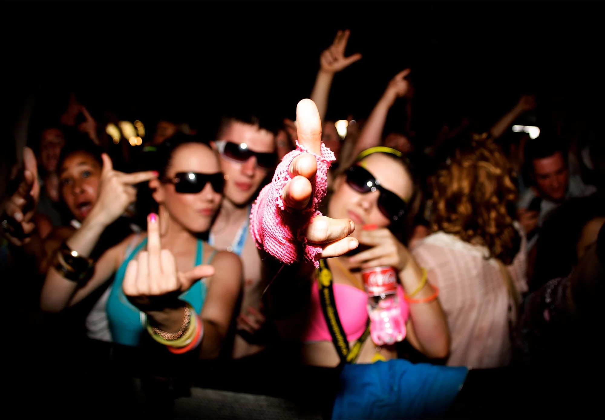 People at a party wearing colorful clothes and sunglasses, with one person in the center making a rude hand gesture and pointing upwards.
