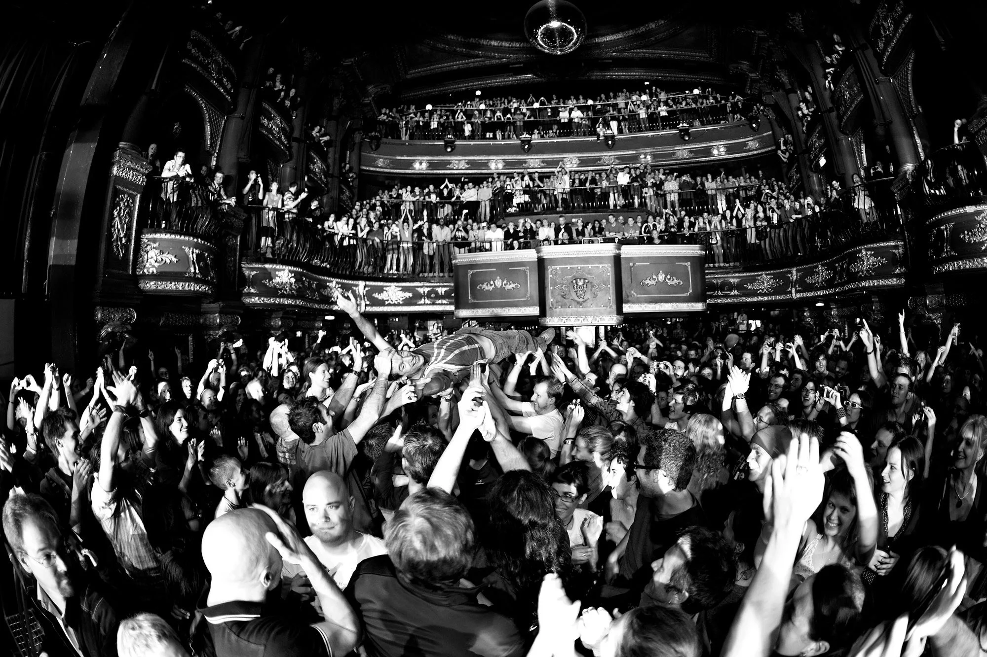 Crowd at a concert or event with multiple levels of balconies filled with people, in a dark indoor venue with ornate details, a central stage, and a person being lifted above the crowd.