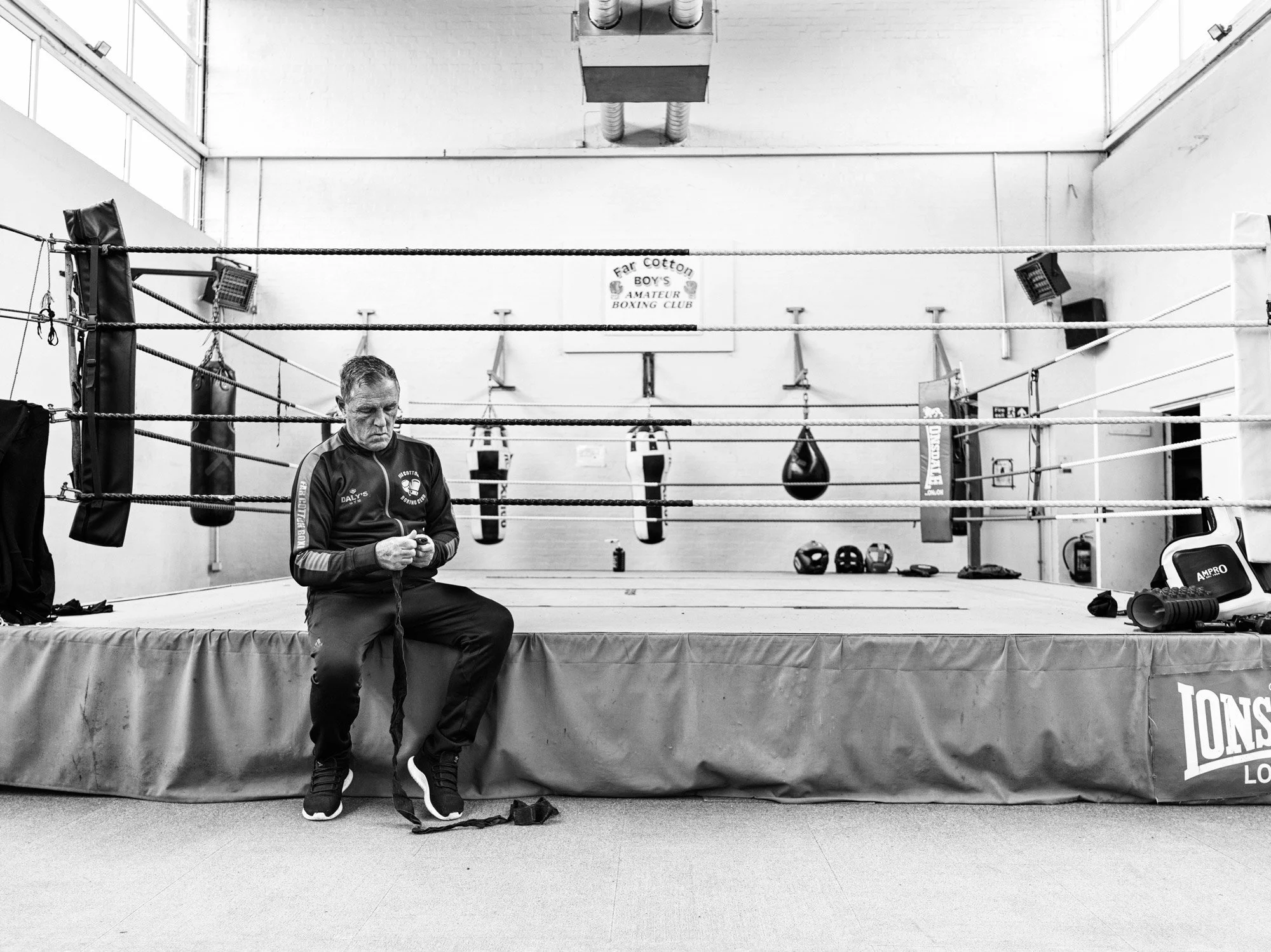 A man sits on the edge of a boxing ring in a boxing gym, looking down at a phone. The gym has boxing punching bags hanging in the background and a sign that reads 'Far Cotton Boys Amateur Boxing Club'.