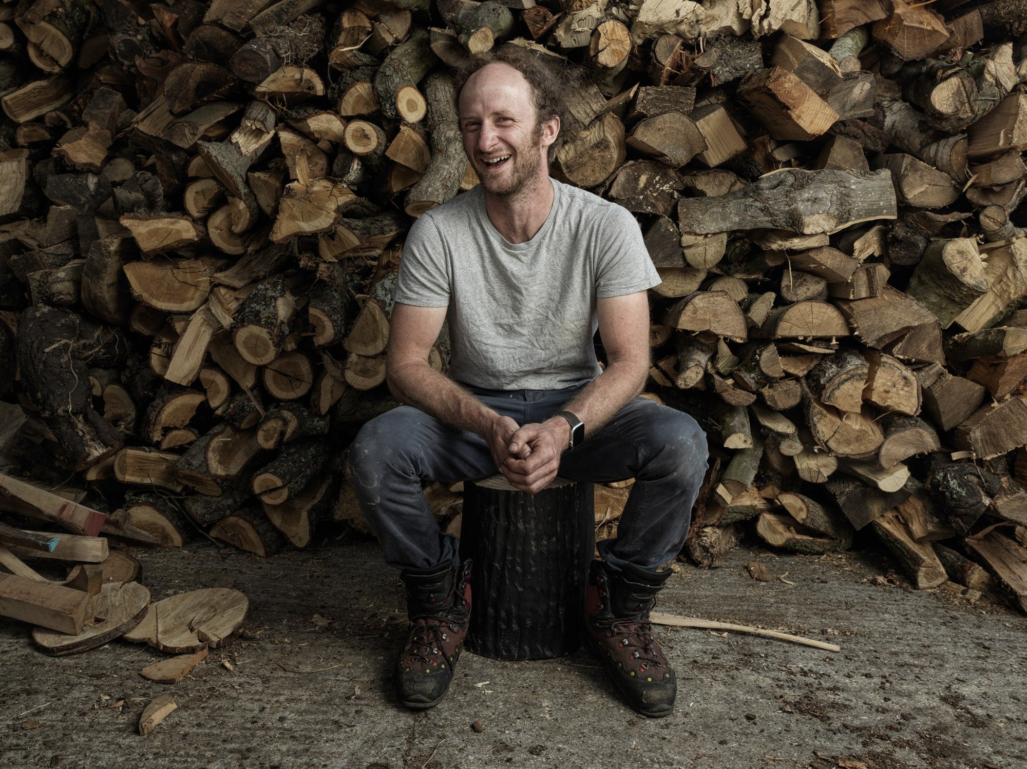 A man sitting on a tree stump in front of a large pile of stacked firewood, smiling and wearing a light gray t-shirt, dark pants, and hiking boots.