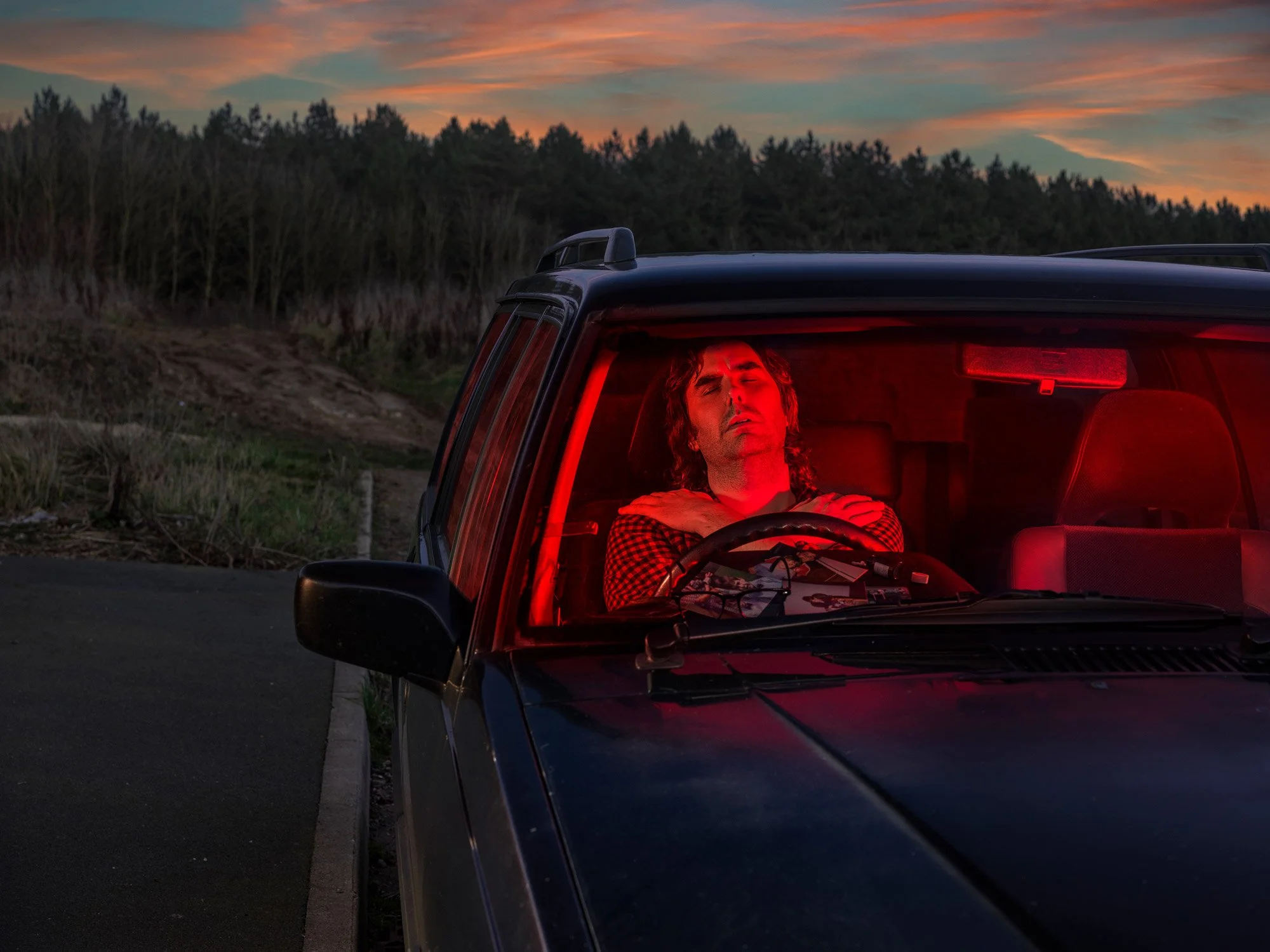 A person with closed eyes and a relaxed expression resting against the back of a car's interior, illuminated by red light, with a sunset sky and trees in the background.