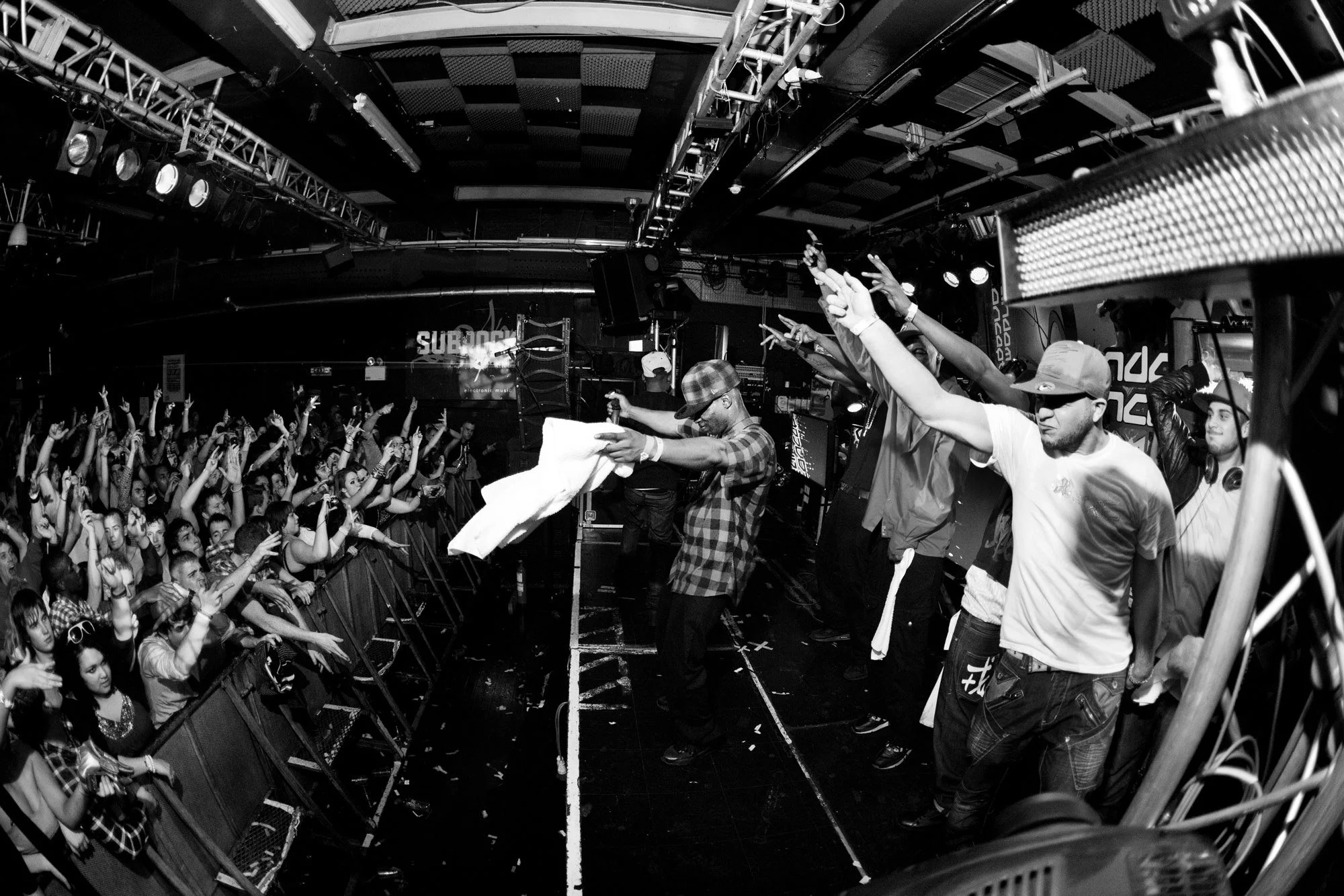 Chase and Status, enjoying the performance with arms raised. The photo shows performers on stage with DJ equipment and stage lights overhead.