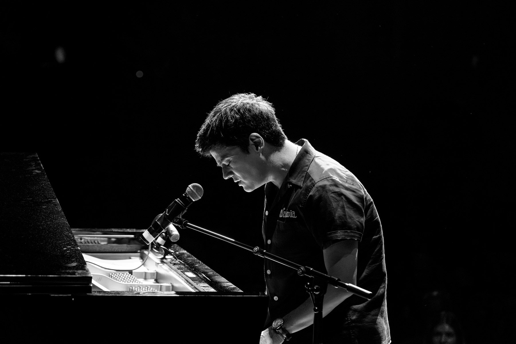 Jamie Cullum playing a piano and singing into a microphone in a dark setting, illuminated by a spotlight.