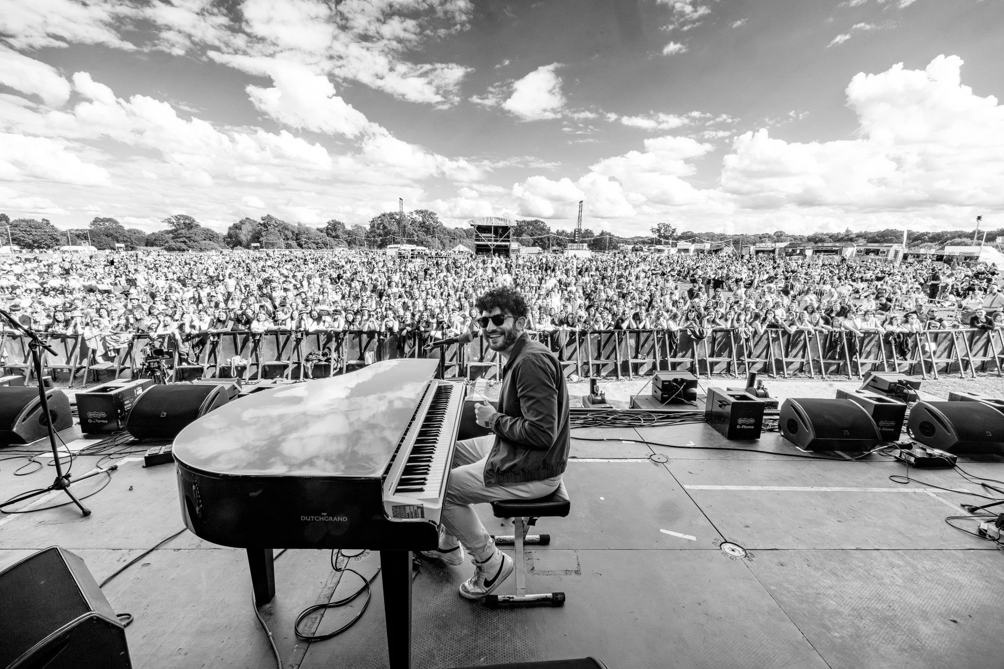 A musician playing a grand piano on an outdoor stage during a large festival with a crowd of people in the background, under a partly cloudy sky.