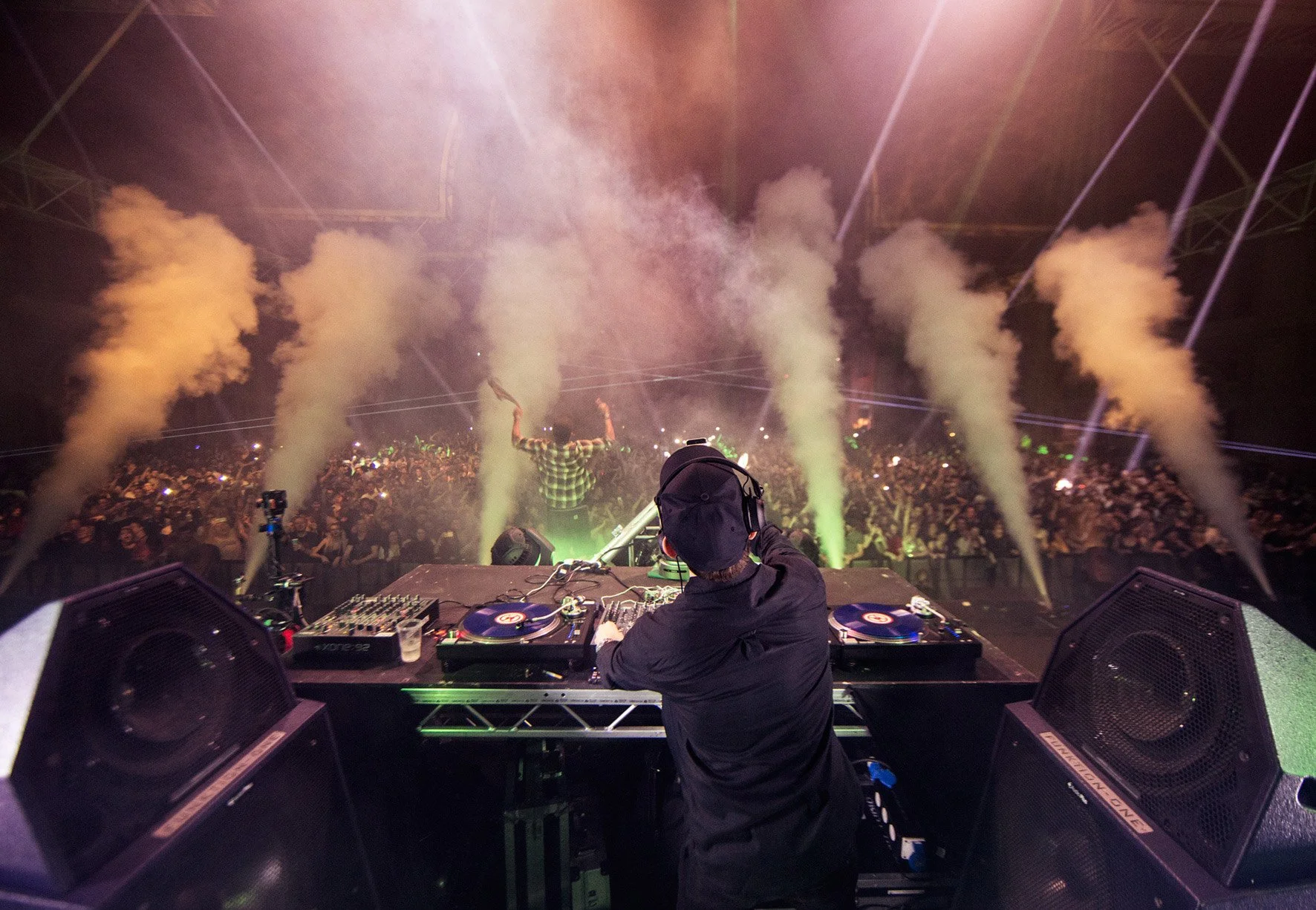 DJ Andy C at a turntable on a stage at Alexandra Palace London, performing for a large crowd at a concert or music event, with smoke and laser lights creating a vibrant atmosphere.