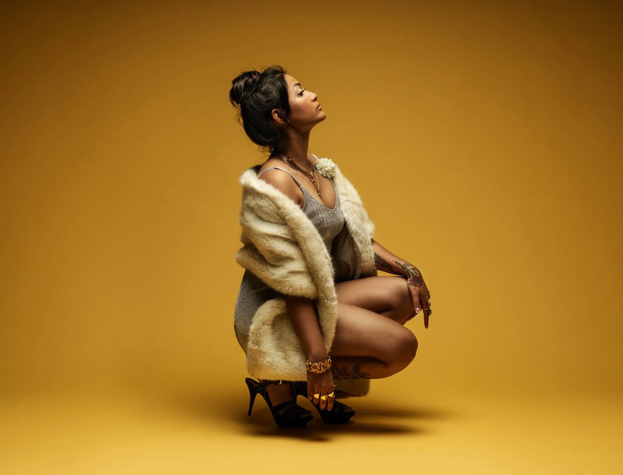 A woman with black hair and tattoos on her left arm poses against a yellow background. She wears a fur coat, lingerie, and high heel shoes, accessorized with jewelry, and is seated with her right leg crossed over her left knee.
