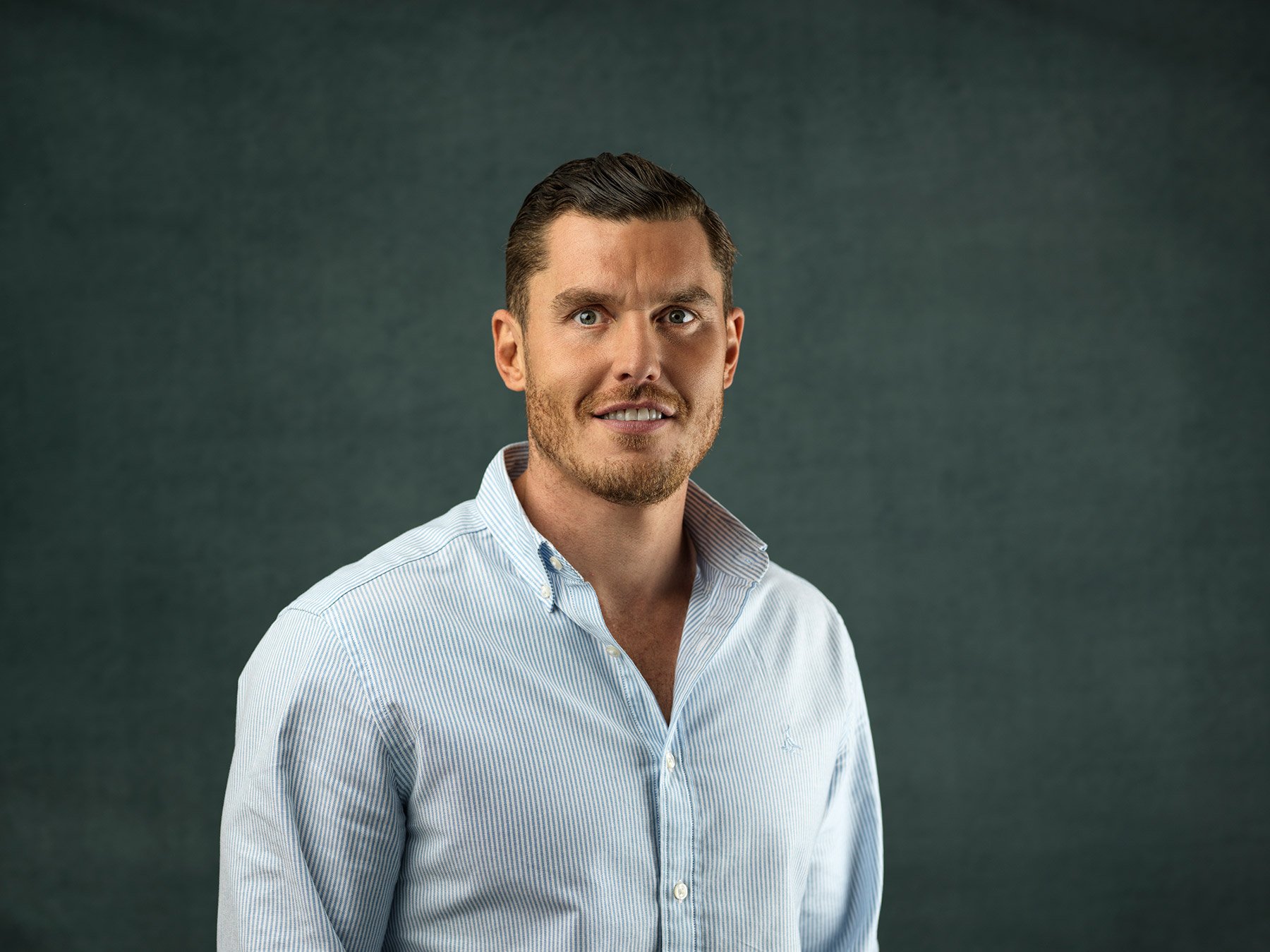 A man with light skin, brown hair, and blue eyes, wearing a light blue button-up shirt, smiling at the camera, standing against a dark green background.
