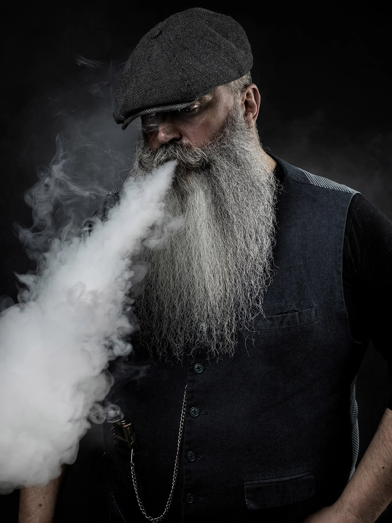 A man with a long gray beard and gray hair, wearing a gray flat cap and a dark vest, is exhaling a large cloud of vapor against a black background.