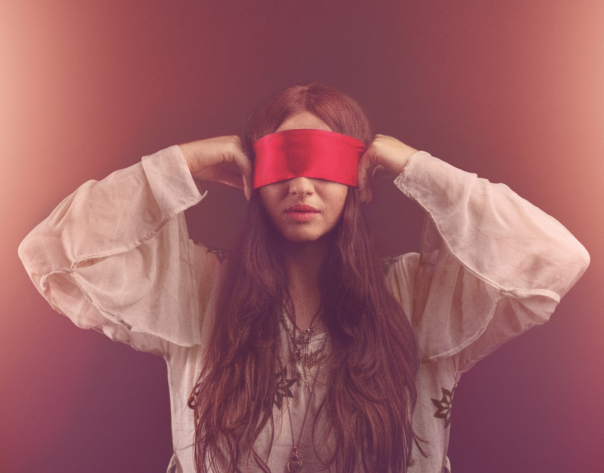 A woman with long, wavy brown hair wearing a white shirt with rolled-up sleeves and a star pattern. She is blindfolded with a red cloth and holding her hands up near her head.