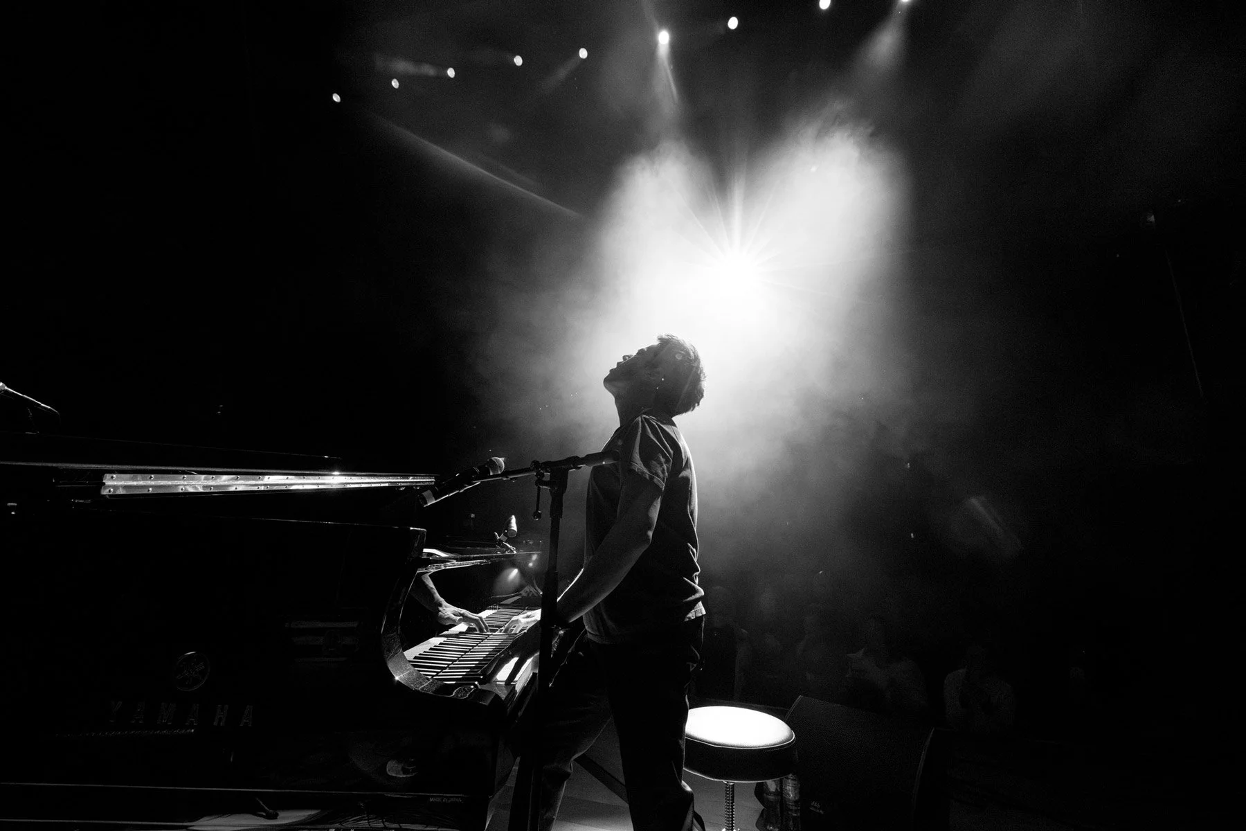 Jamie Cullum playing piano on stage in black and white, with dramatic backlighting creating a silhouette and spotlight effects.