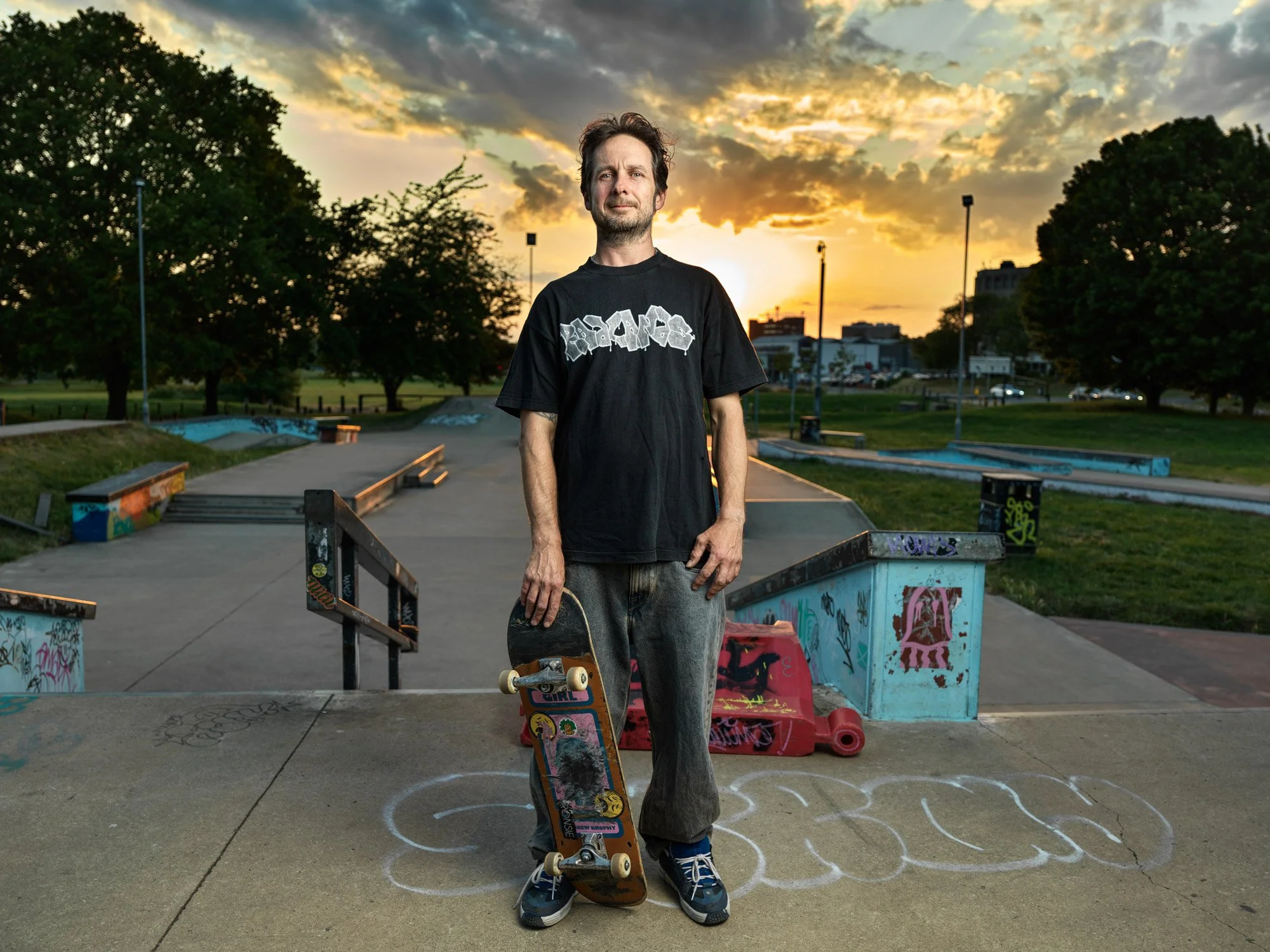 A man standing with a skateboard at a skate park during sunset. He is wearing a black t-shirt with graffiti-style text, gray pants, and blue sneakers. The skate park has graffiti-covered rails and ramps, and the sky is partly cloudy with the sun sett