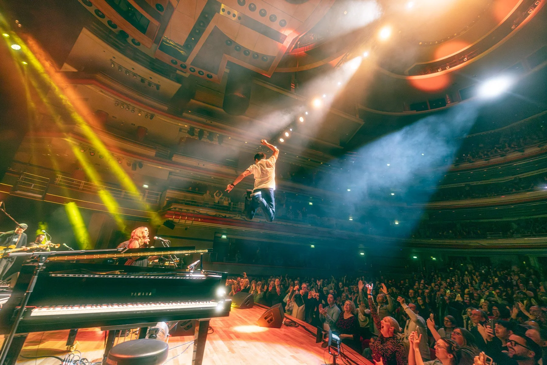 A performer jumps in mid-air on stage during a concert at a theater, with bright stage lights and a cheering audience. Musicians and instruments are visible, including a person playing a keyboard in the foreground.
