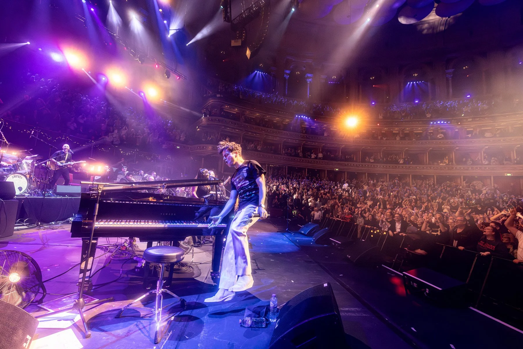 A lively concert scene in a theater with a stage and purple and yellow lighting. A musician is adjusting a grand piano in the foreground, while other band members play guitars and drums on stage. The audience fills the theater, with some standing and