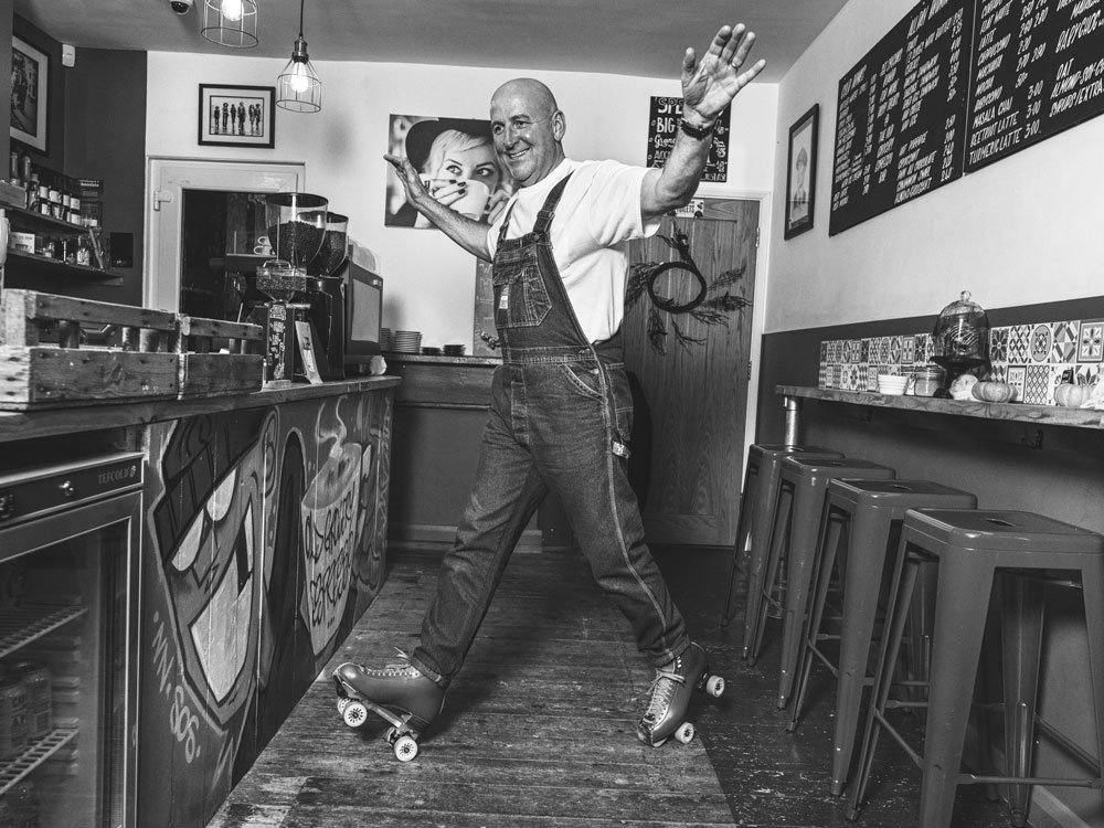 A man roller skating inside a cafe, smiling and waving at the camera.
