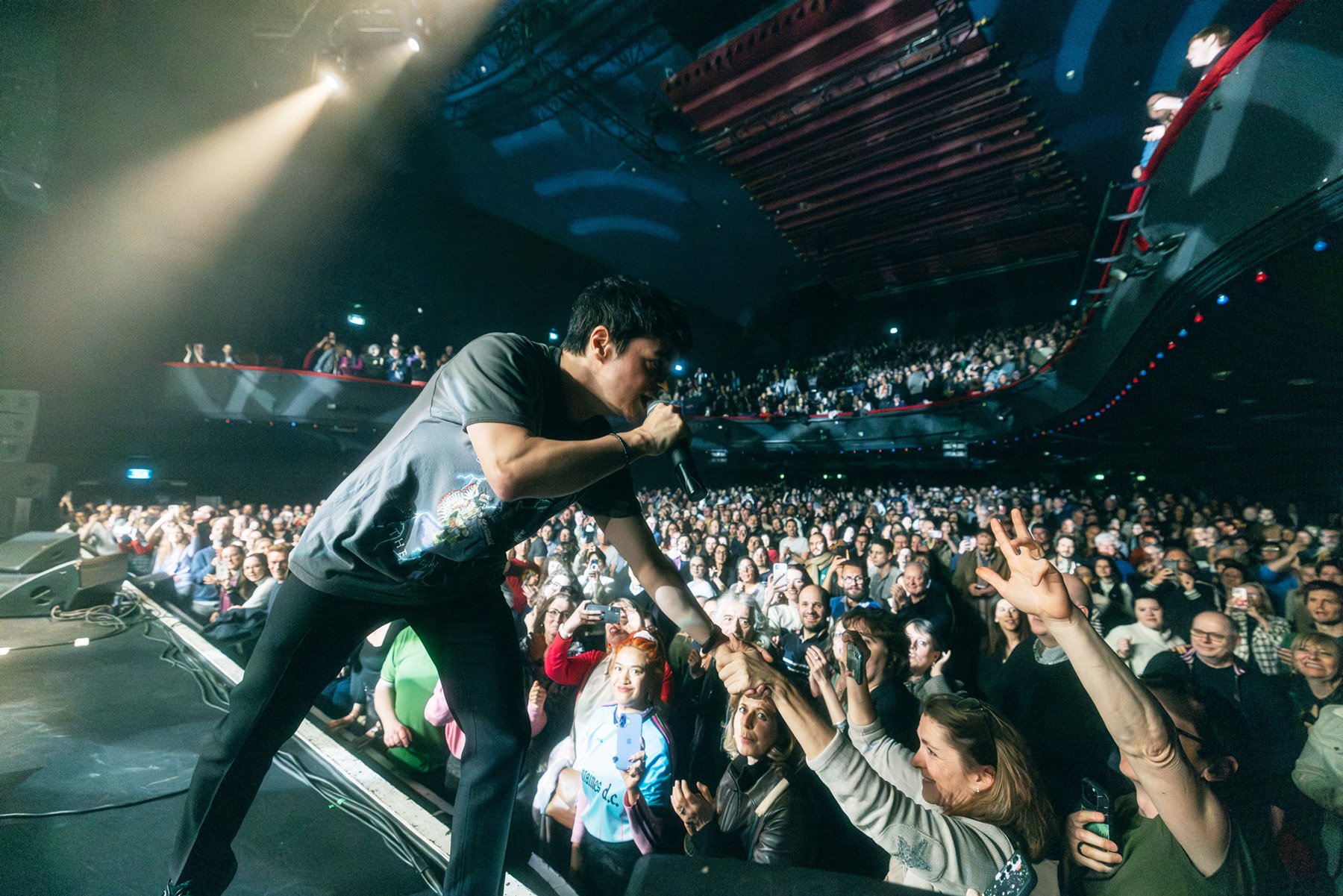 Jamie Cullum performs on stage while reaching out to an enthusiastic audience, with fans showing excitement and taking photos and videos during a concert in a packed venue.