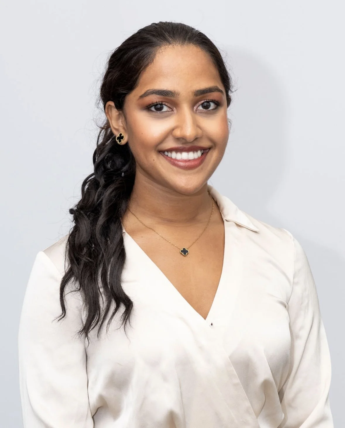 A woman with dark hair styled in loose waves, wearing a white blouse, gold and black jewelry, smiling against a plain light background.