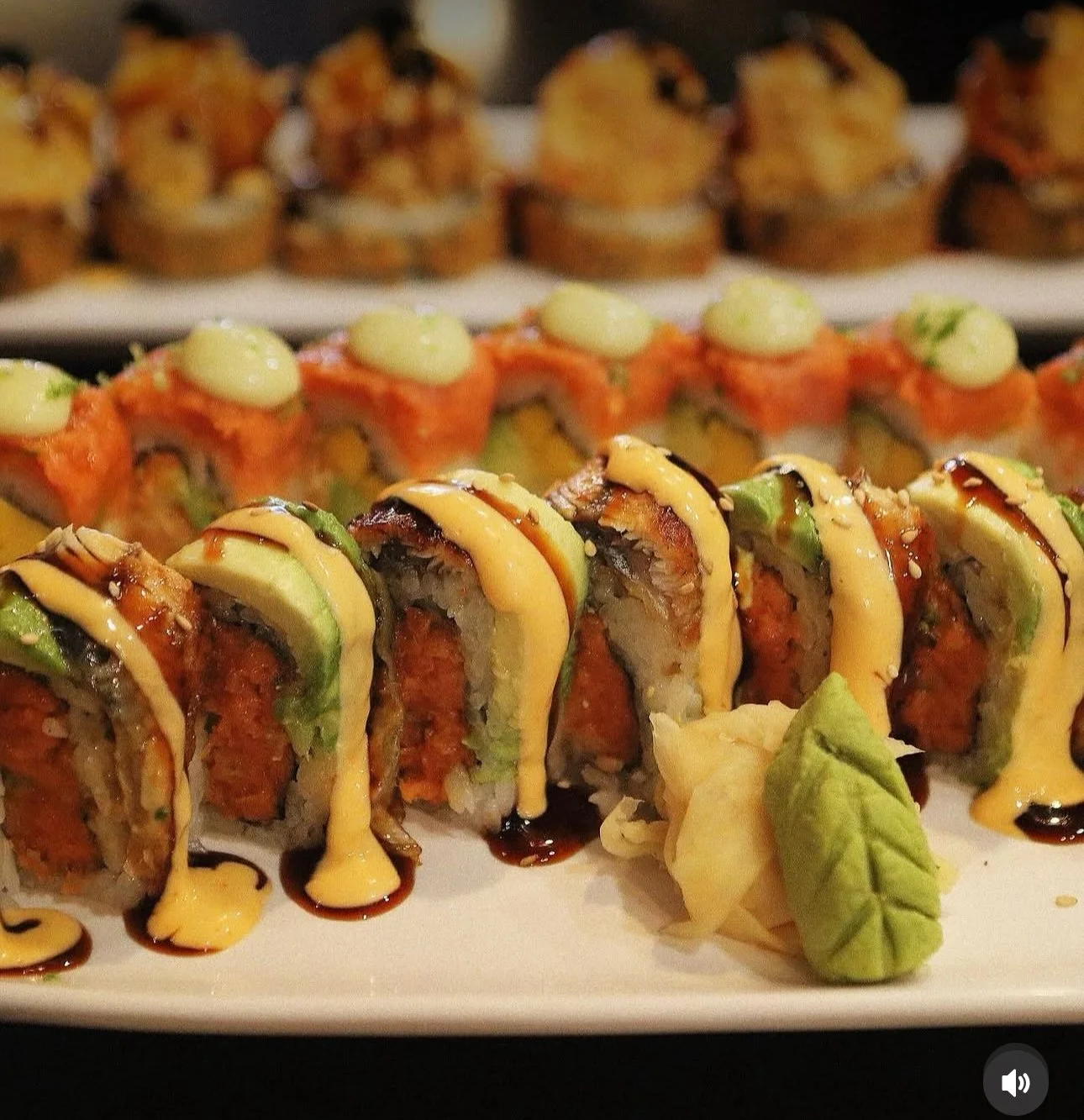 Close-up of assorted sushi rolls topped with sauces, inside a white serving platter, with slices of wasabi and ginger.