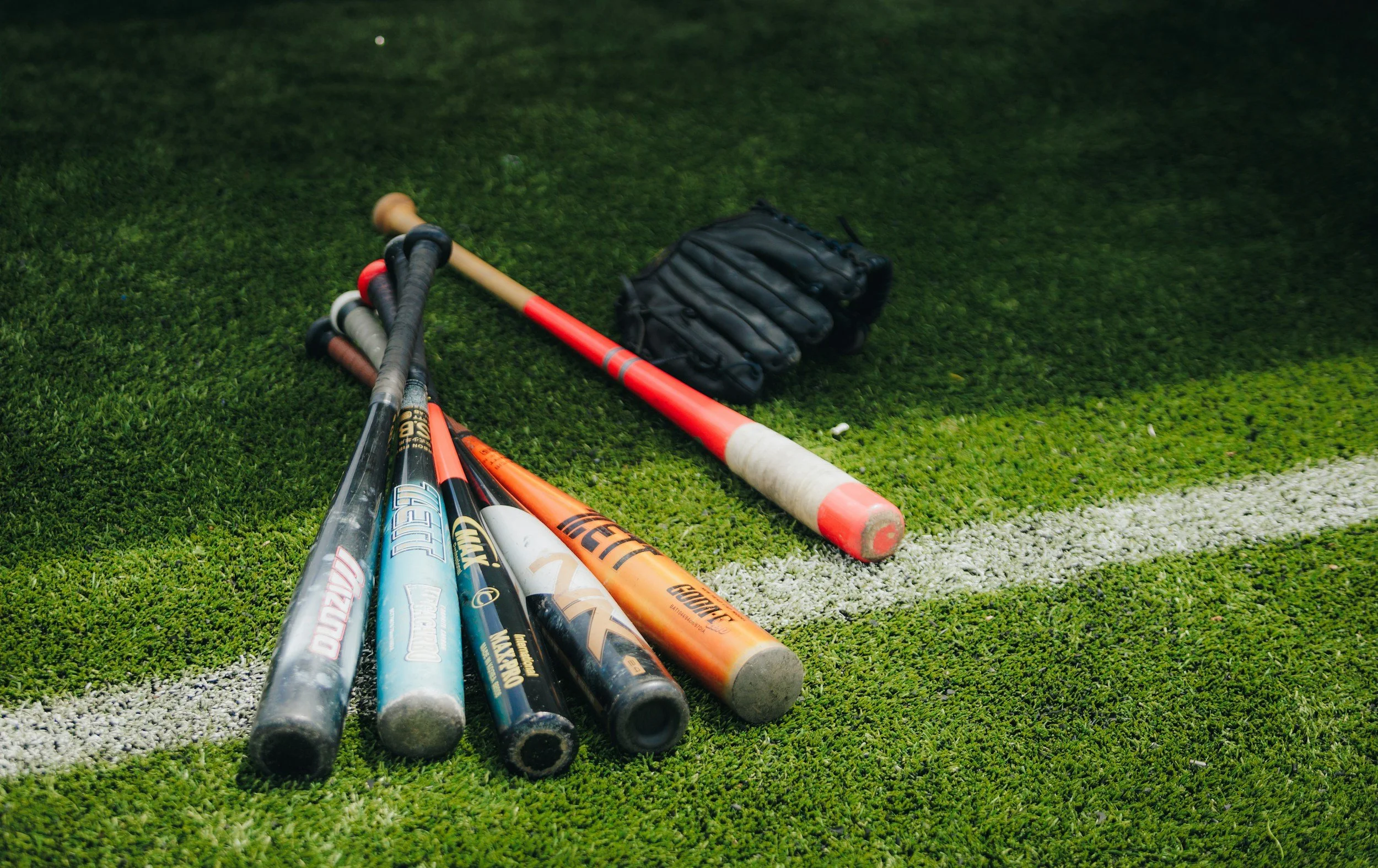 A collection of baseball bats lying on a green artificial turf field, with a black baseball glove nearby.