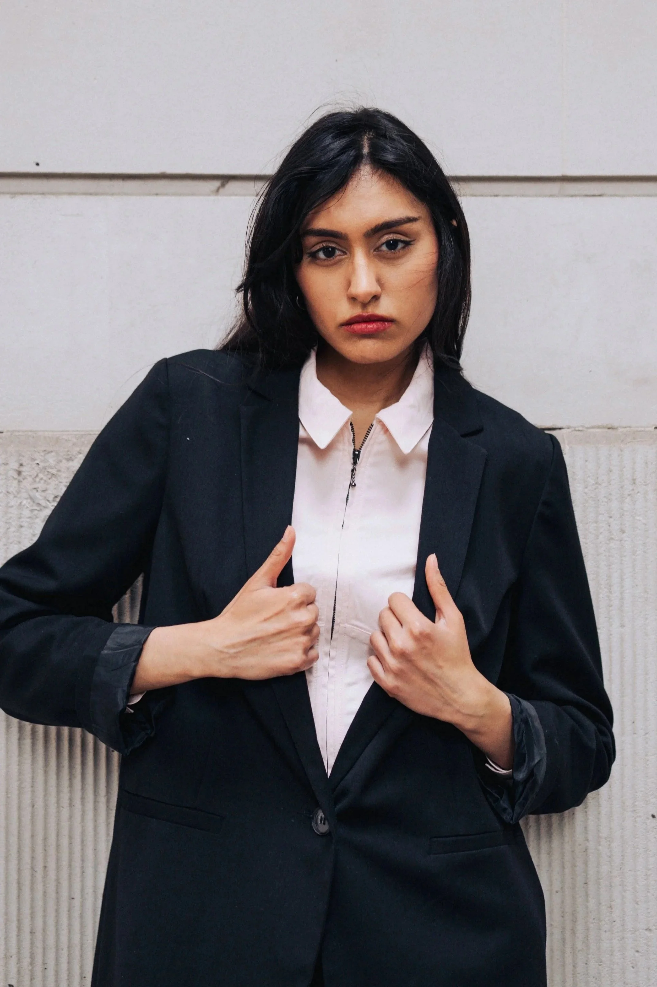 A woman with dark hair wearing a black blazer and white shirt, standing against a ledge wall.