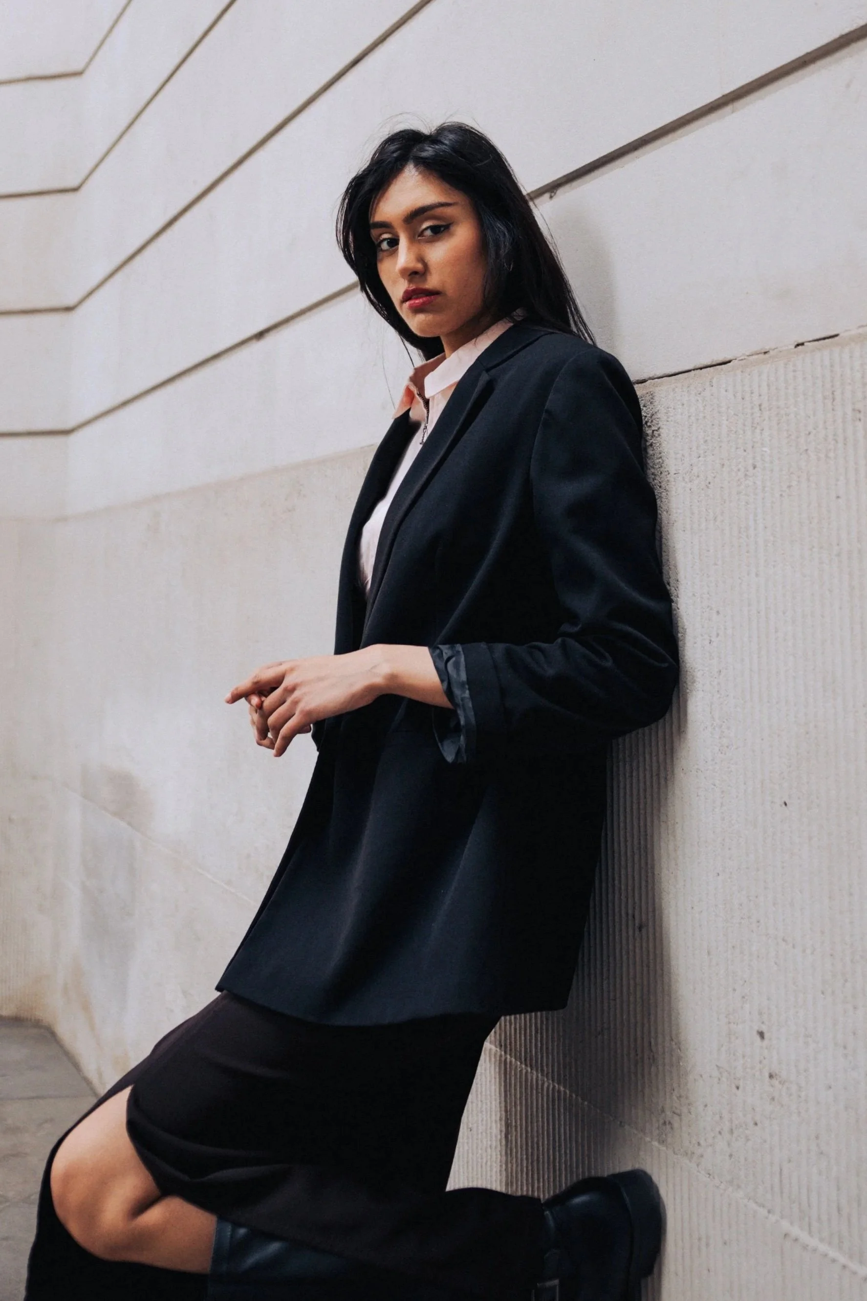 A young woman with dark hair and medium skin tone leaning against a beige wall with horizontal and vertical textured panels, wearing a black blazer over a light pink top, a black skirt, and black boots.