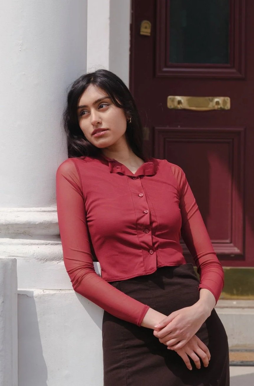 A young woman with dark hair and light skin leaning against a white wall outside a building, wearing a red sheer long-sleeve blouse and a dark skirt, looking contemplative.