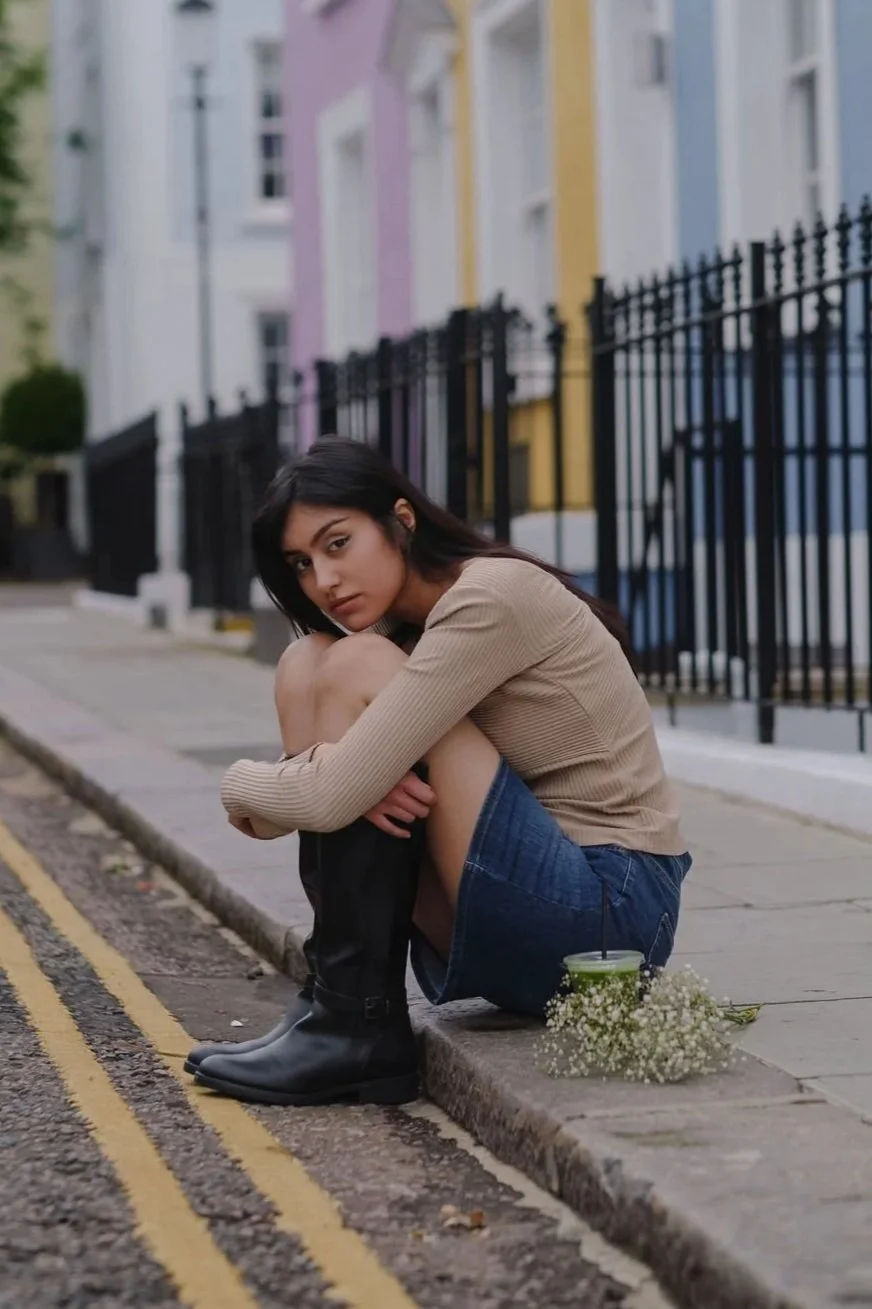 A young woman sitting on a city sidewalk with her knees drawn up, wearing a beige sweater, denim shorts, and black rain boots. She has dark hair and is gazing at the camera with a serious expression. In front of her, there is a small bouquet of white