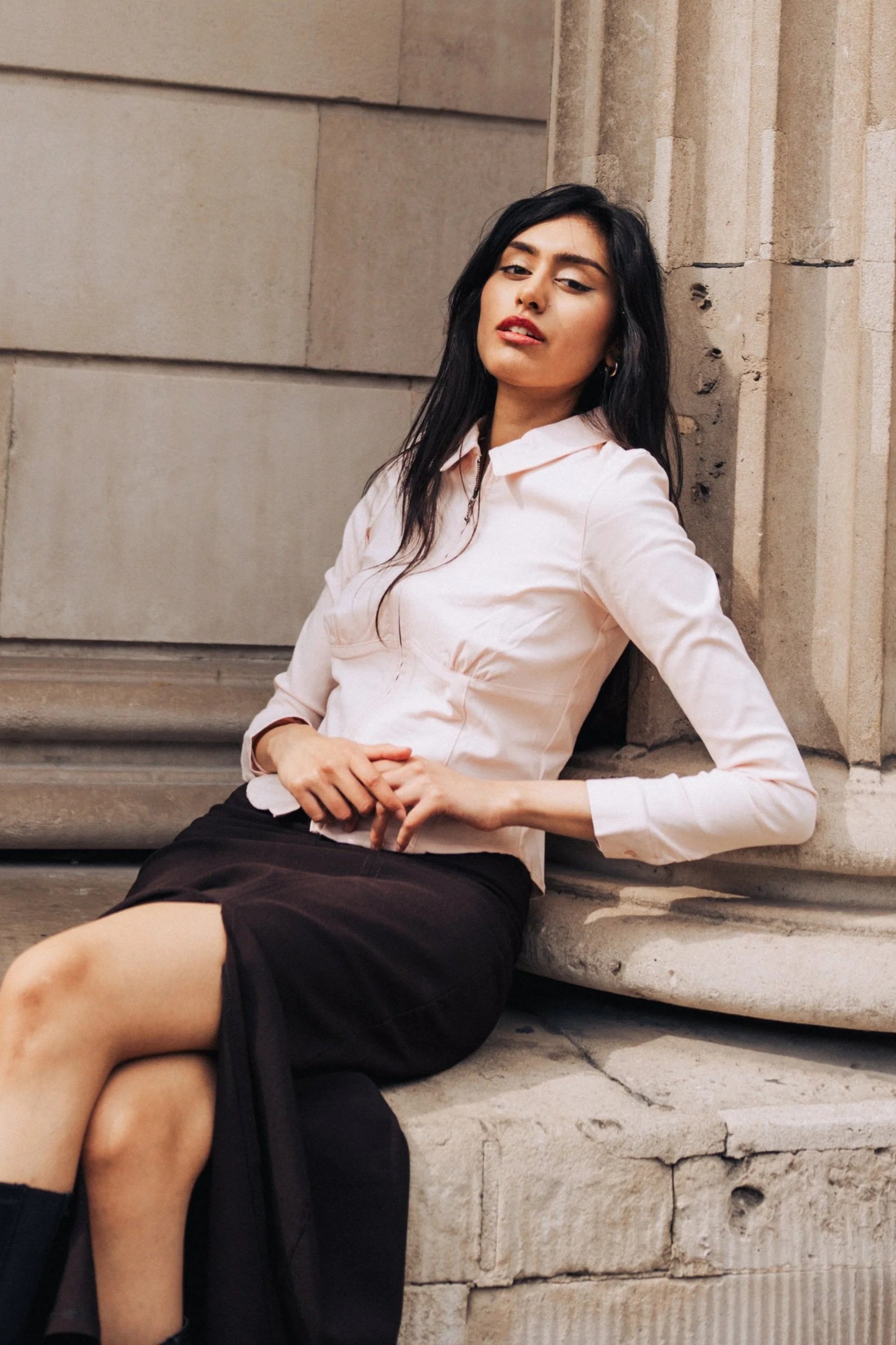 A young woman sits on stone steps next to a stone wall, leaning back with one arm resting on the ledge. She has long dark hair, wears a light-colored blouse and a dark skirt, and looks towards the camera with a relaxed expression.
