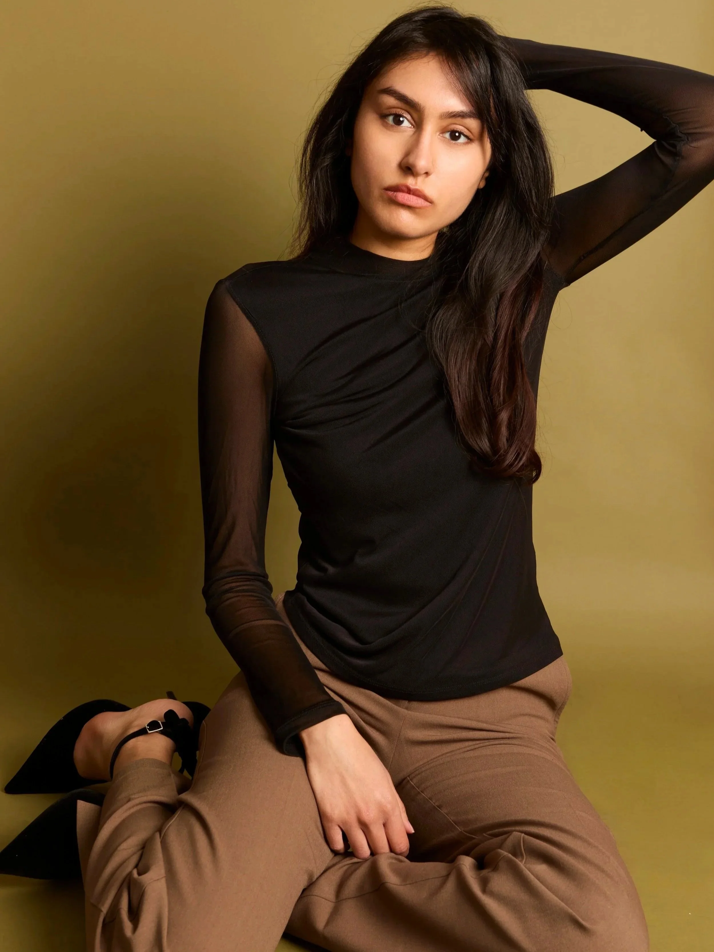 A young woman with long dark hair wearing a black sheer long-sleeve top and beige pants, seated on the floor against a yellow background, with her arm raised behind her head.