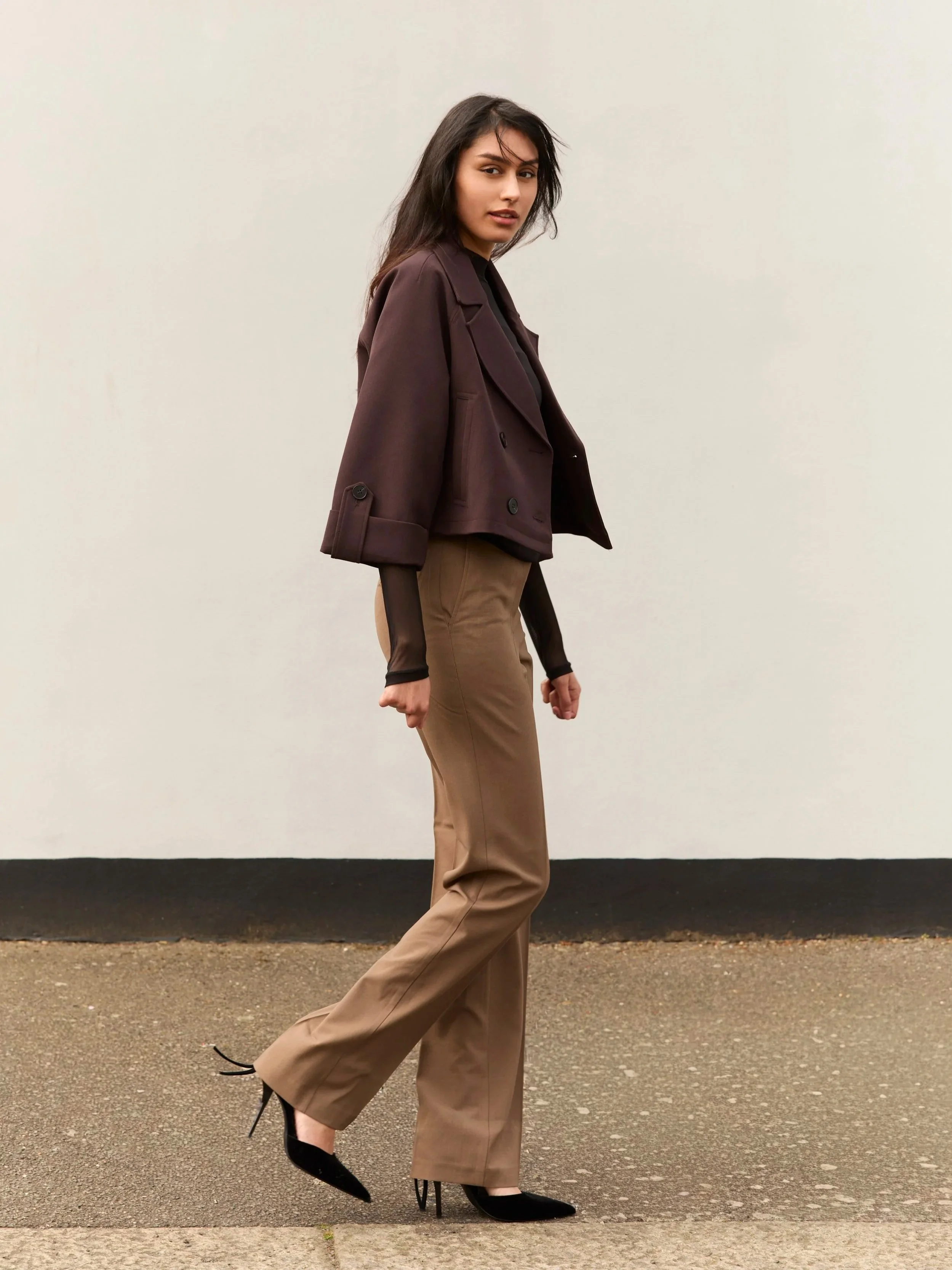 A woman with long dark hair wearing a brown blazer, black sheer top, beige pants, and black high heels walking outdoors on a paved surface against a plain, light-colored wall.