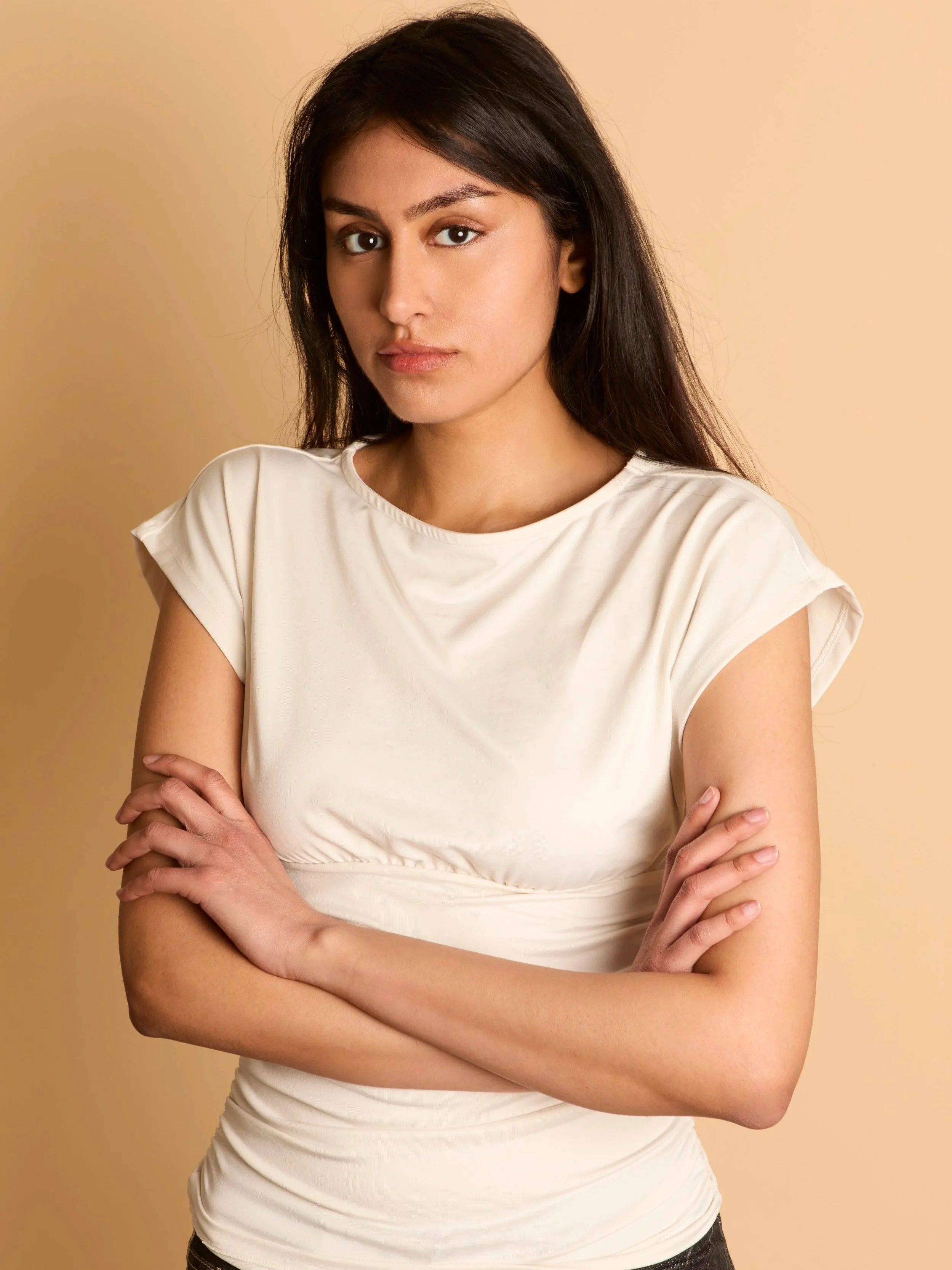 A woman with dark brown hair and light skin stands with her arms crossed against a beige background, wearing a cream-colored top.