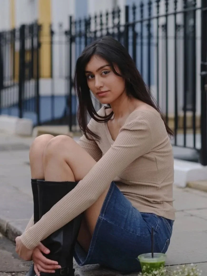Young woman sitting on the sidewalk near a fence, holding her knee, with a green drink nearby.