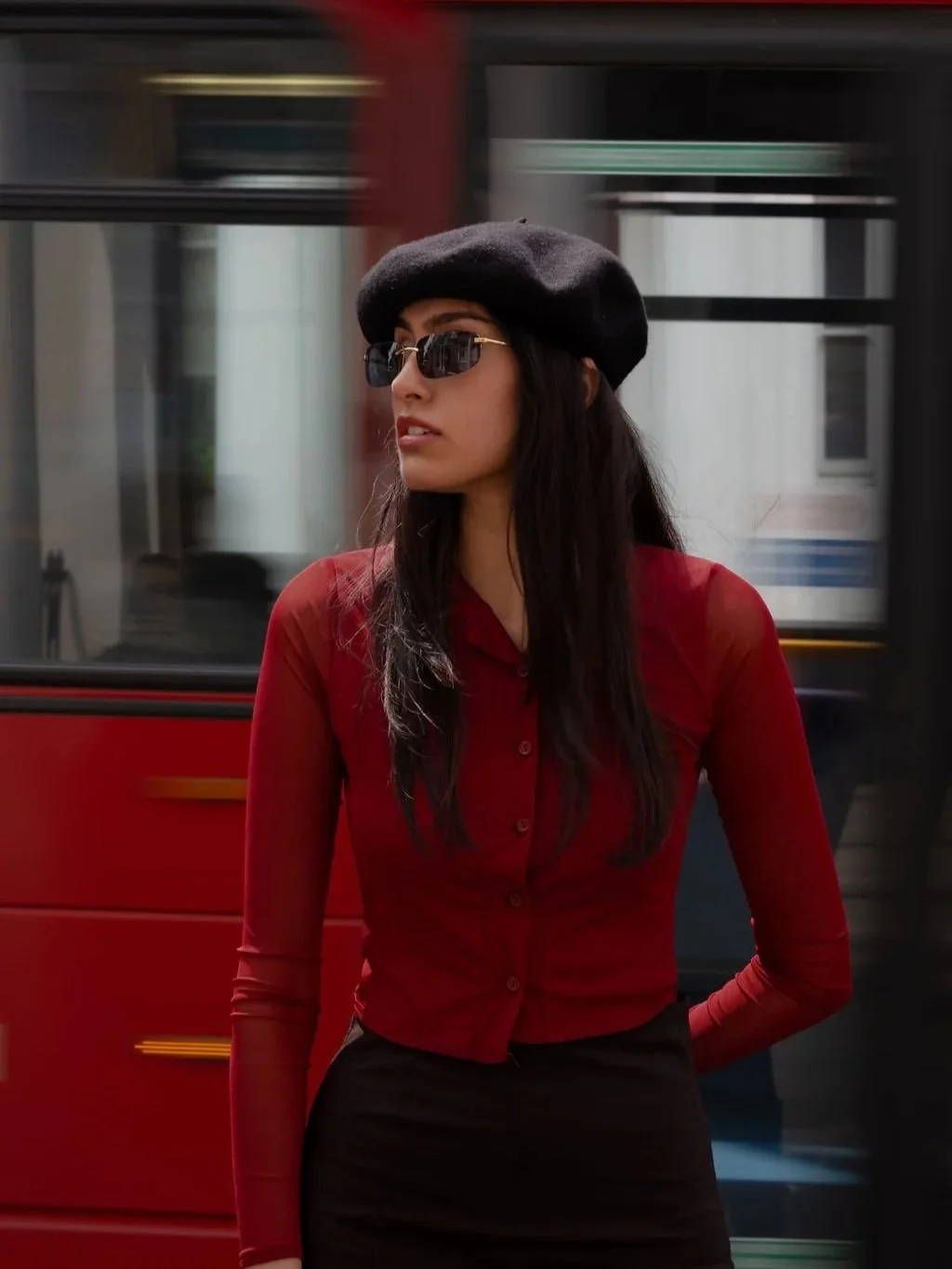 Woman with long dark hair wearing sunglasses, a black beret, a red button-up top, and black skirt walking outdoors with a moving bus in the background.