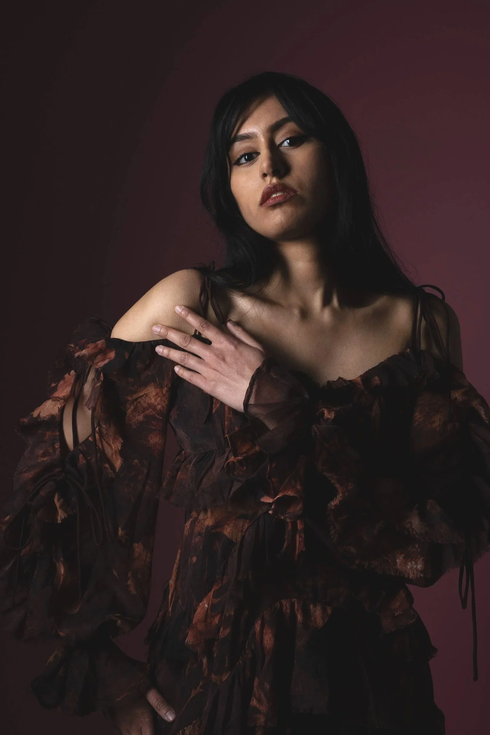 A woman with long dark hair wearing a dark floral off-shoulder dress poses against a dark background, gazing directly at the camera with her hand on her shoulder.