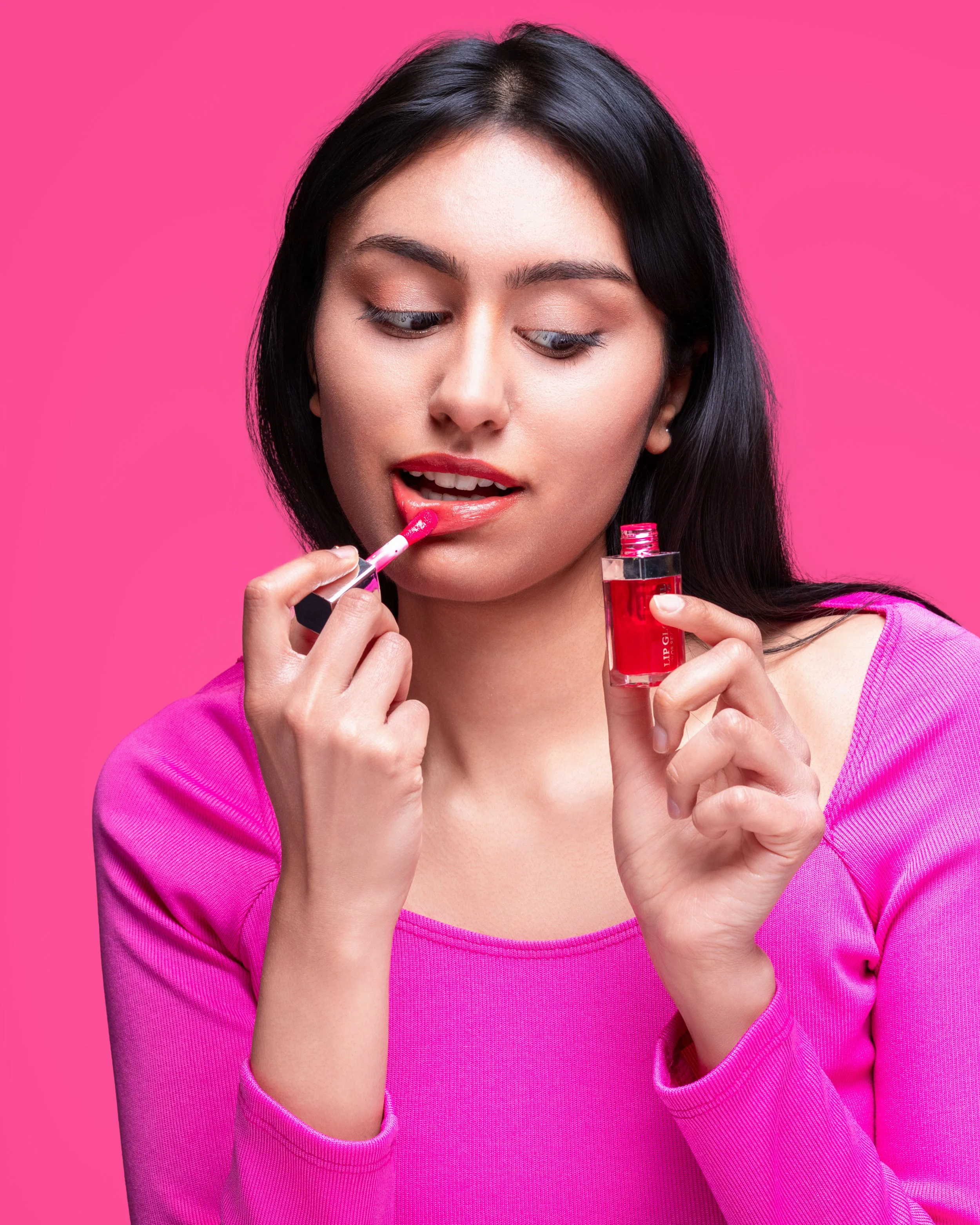 Woman with long black hair applying red lipstick, wearing a pink top, against a pink background.