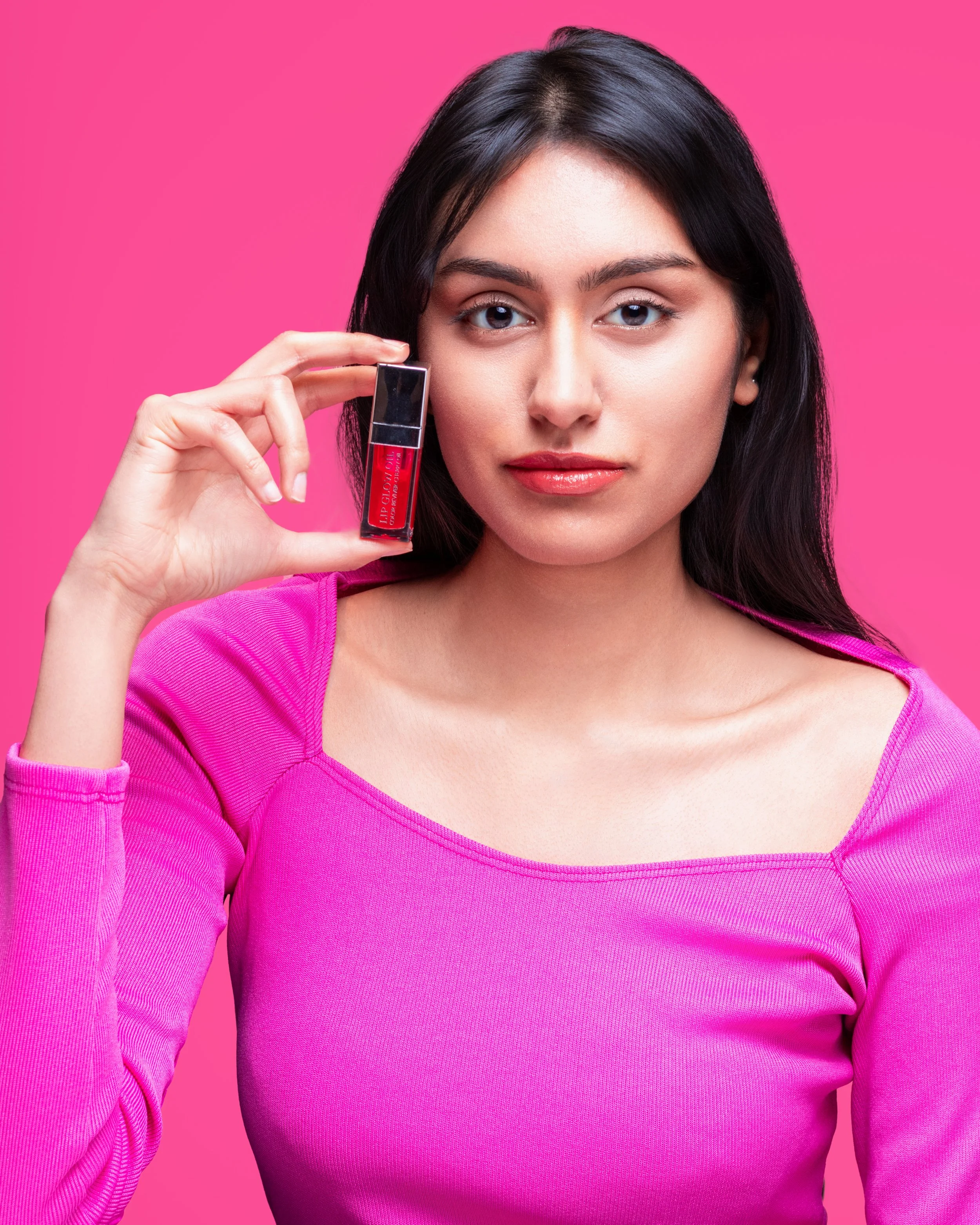 A woman with long dark hair and light skin looking at the camera against a pink background, holding a tube of red lipstick near her face.