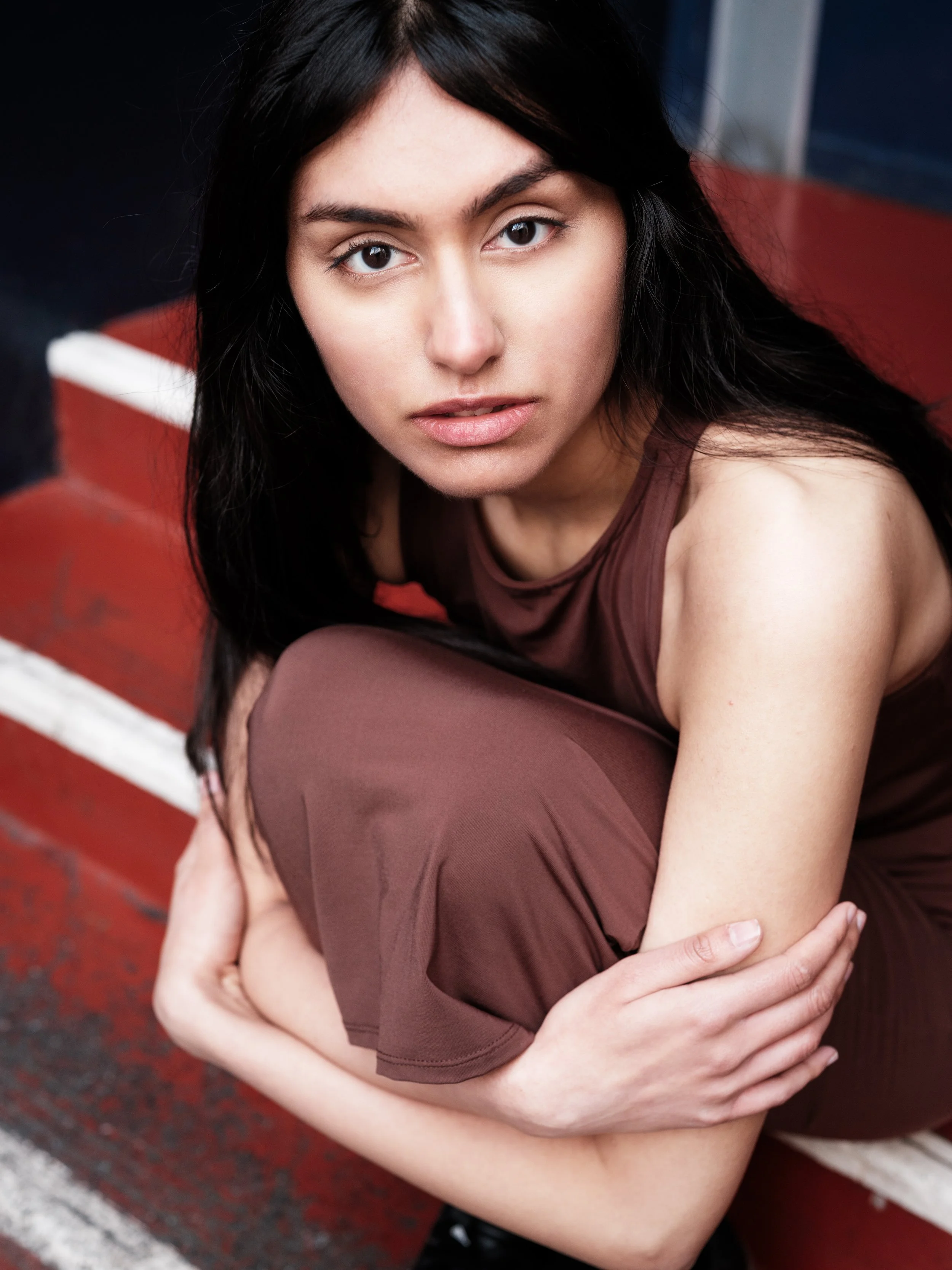 A young woman with long black hair looking directly at the camera with a serious expression, sitting on a red running track with white lines.
