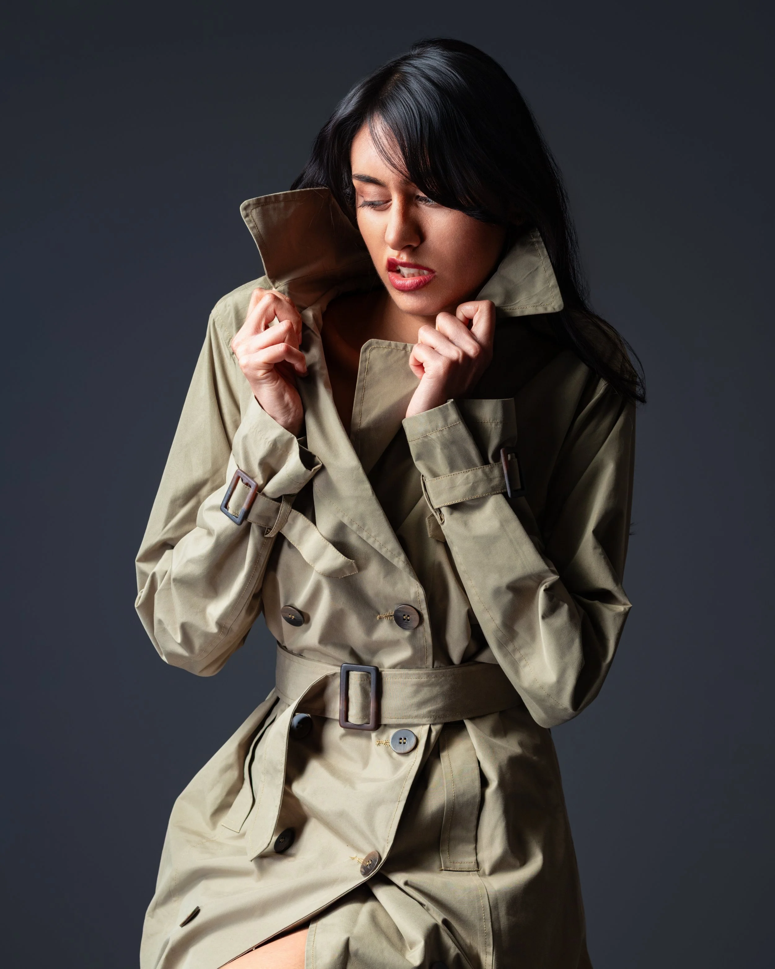A woman with dark hair wearing a beige trench coat holding the collar close to her face, against a dark background.