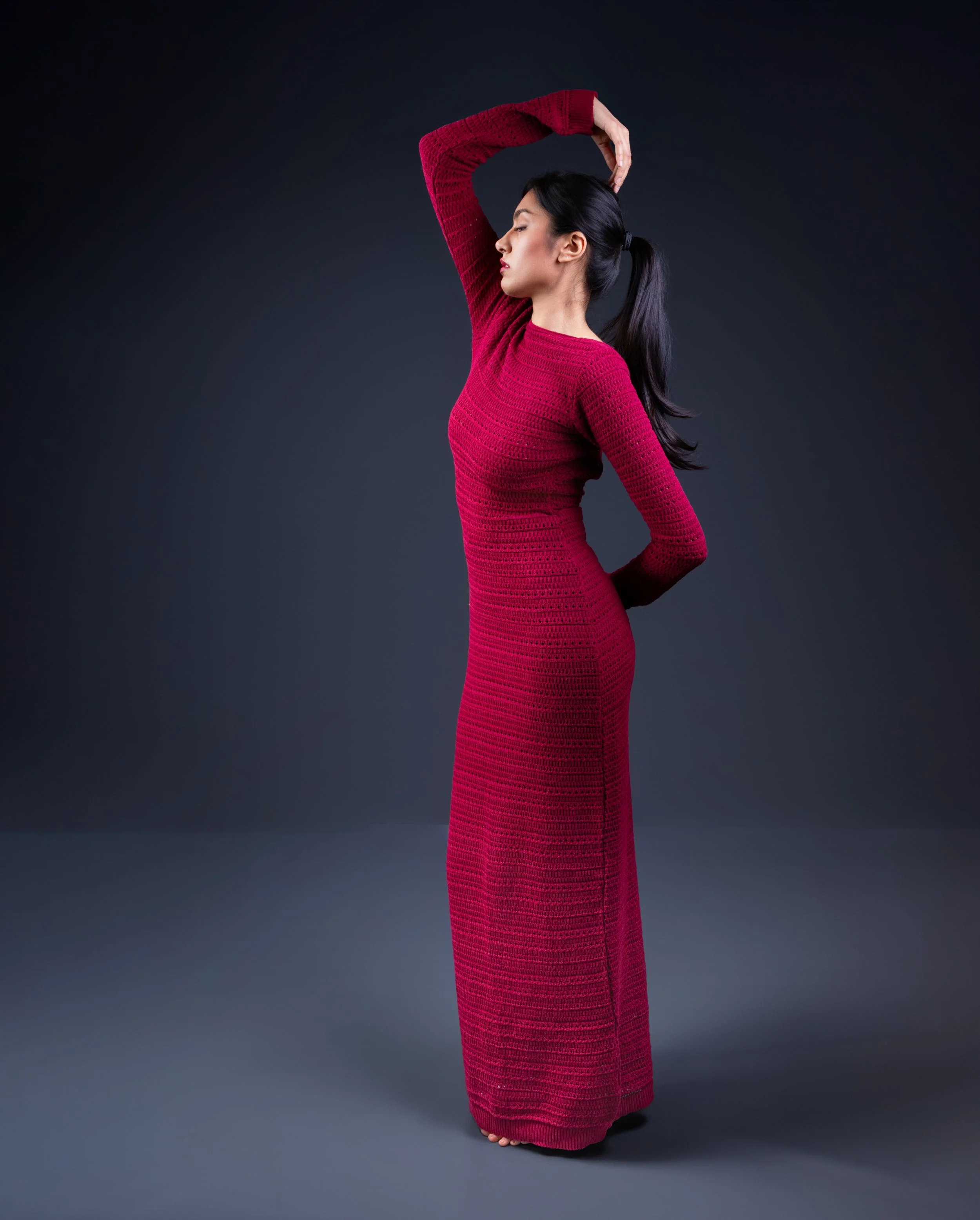 A woman with long dark hair in a high ponytail stands sideways with her eyes closed, wearing a long, fitted red knit dress and raising her left arm above her head against a dark background.